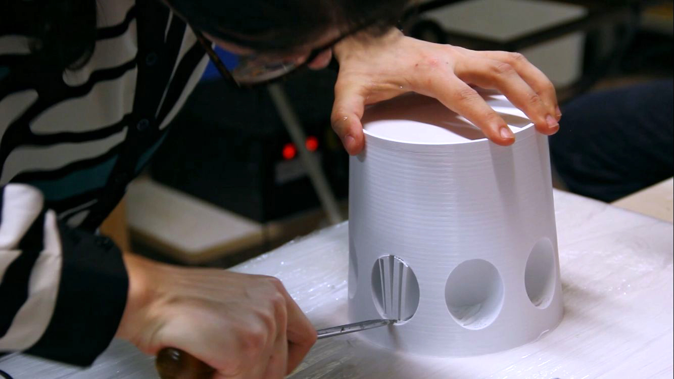 Artigiano che scolpisce fori su un contenitore cilindrico bianco con un coltello su un tavolo di lavoro.