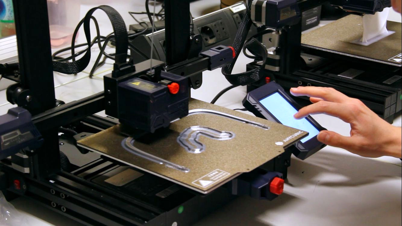 Stampante 3D in funzione su banco di lavoro con uno schermo touchscreen e mano che interagisce.