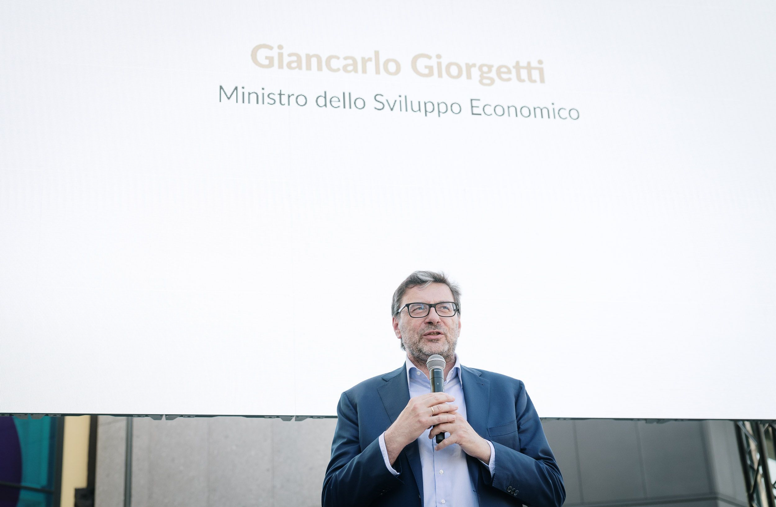 Una persona in abito formale parla al microfono su un palco, con sfondo bianco e testo sopra di lui.