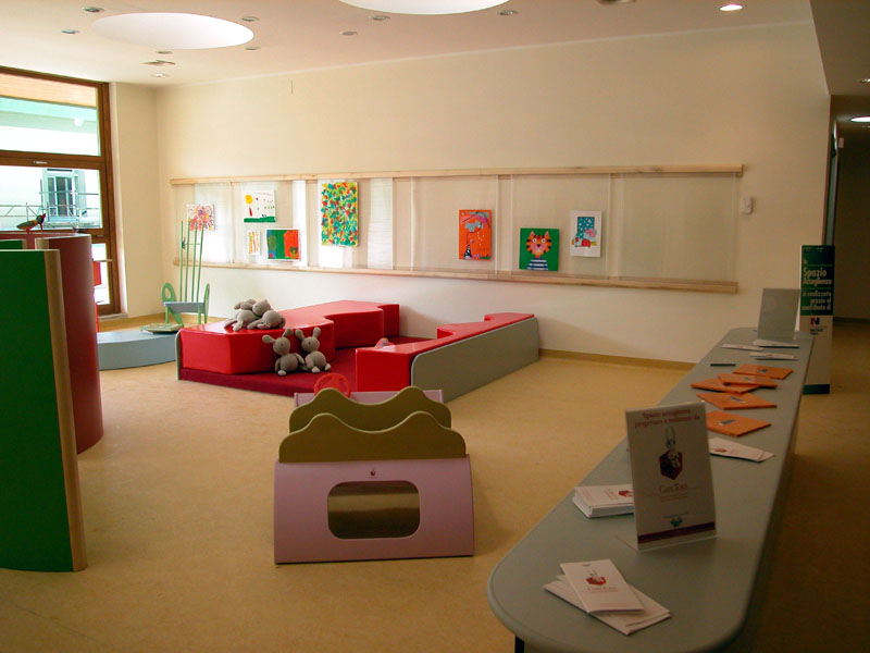 Sala giochi luminosa con arredi colorati e peluche, tavolo laterale con brochure e arte appesa alle pareti.
