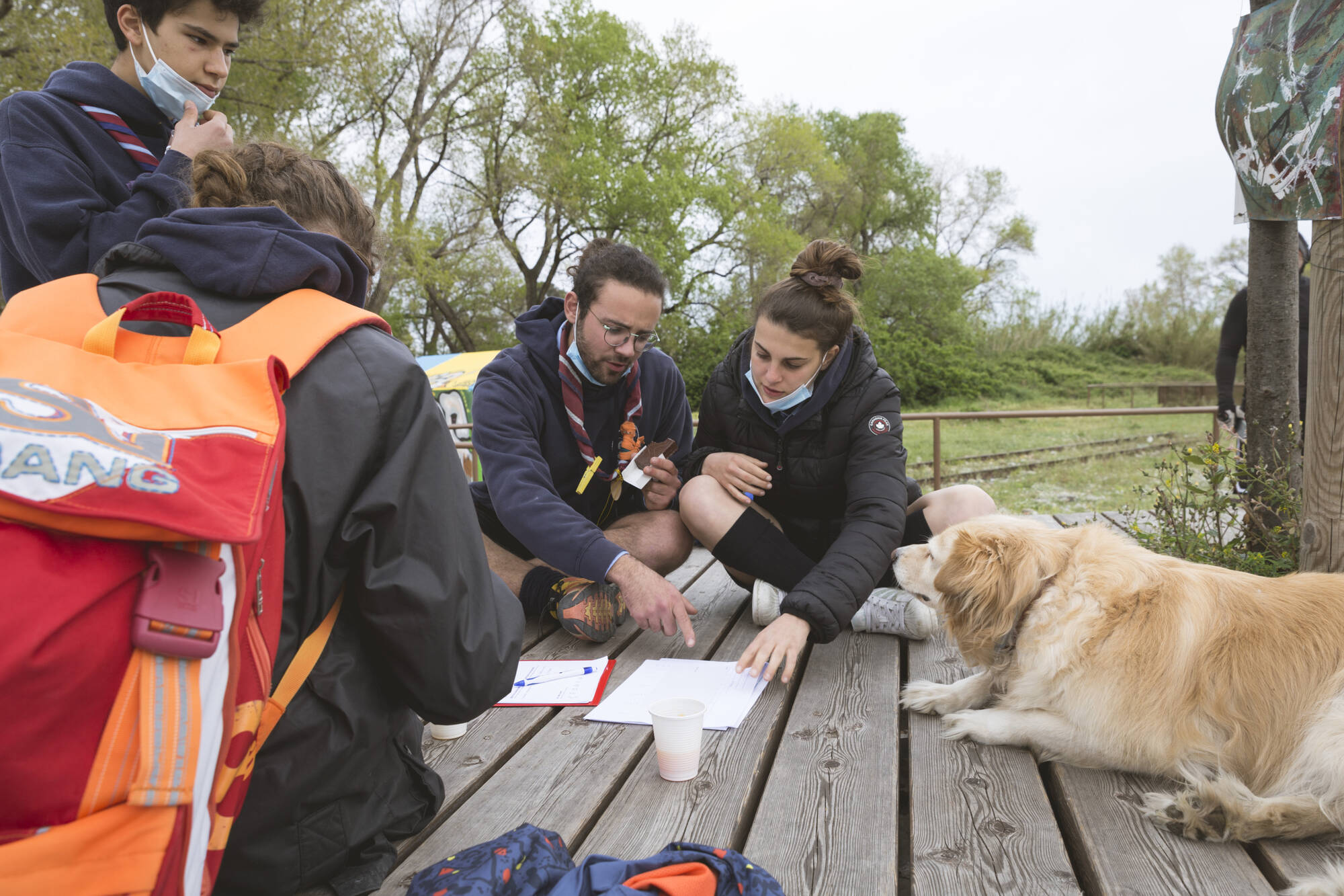 Giovani scout studiano mappe su una piattaforma di legno all'aperto, con un cane che li osserva attentamente.