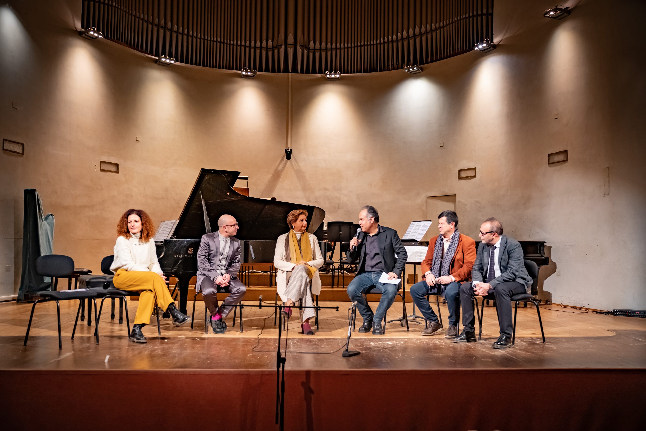 Esperti in un teatro si confrontano davanti a un pianoforte; atmosfera di discussione e cultura.