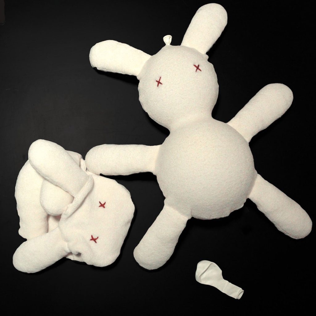 Peluches bianchi a forma di conigli strappati su sfondo nero, con piccole croci rosse sugli occhi.