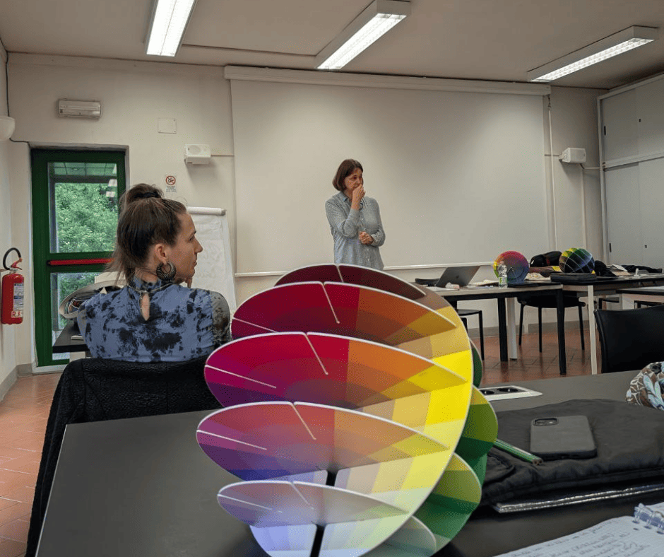 Aula con due persone, una seduta e una in piedi, tavolo con oggetti colorati e uno schema cromatico in primo piano.