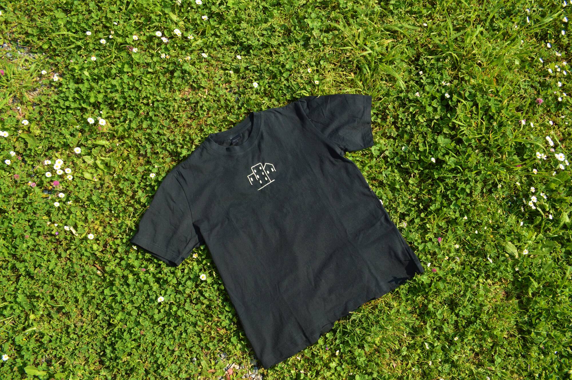 Maglietta nera con un piccolo disegno bianco, stesa su prato verde con fiori bianchi e viola.