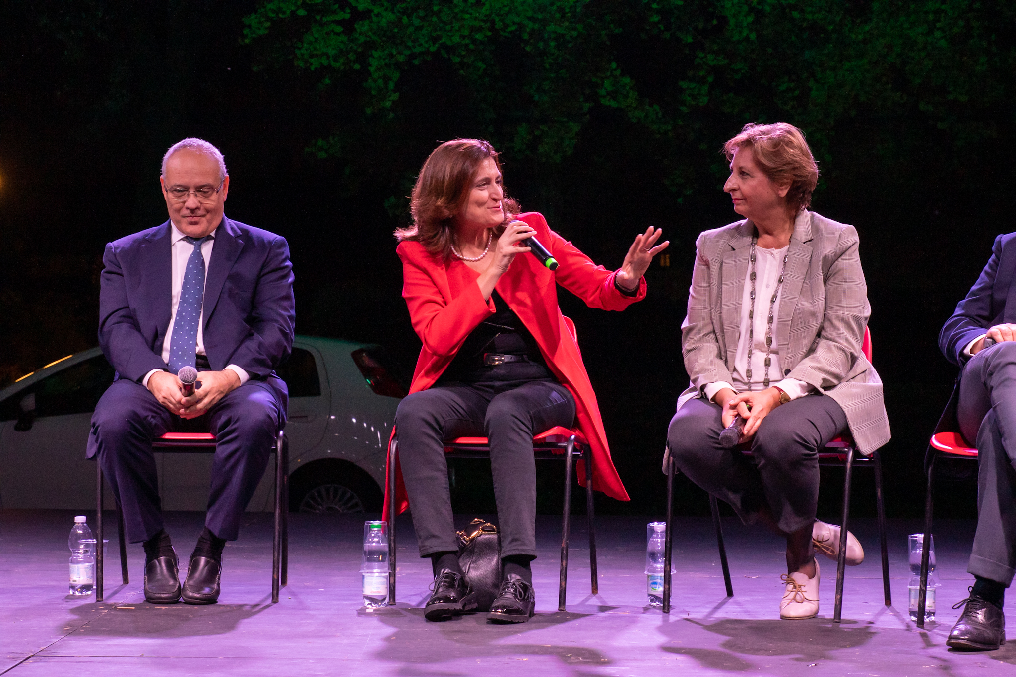 Tre persone sedute su un palco in un dibattito pubblico, una donna parla al microfono.