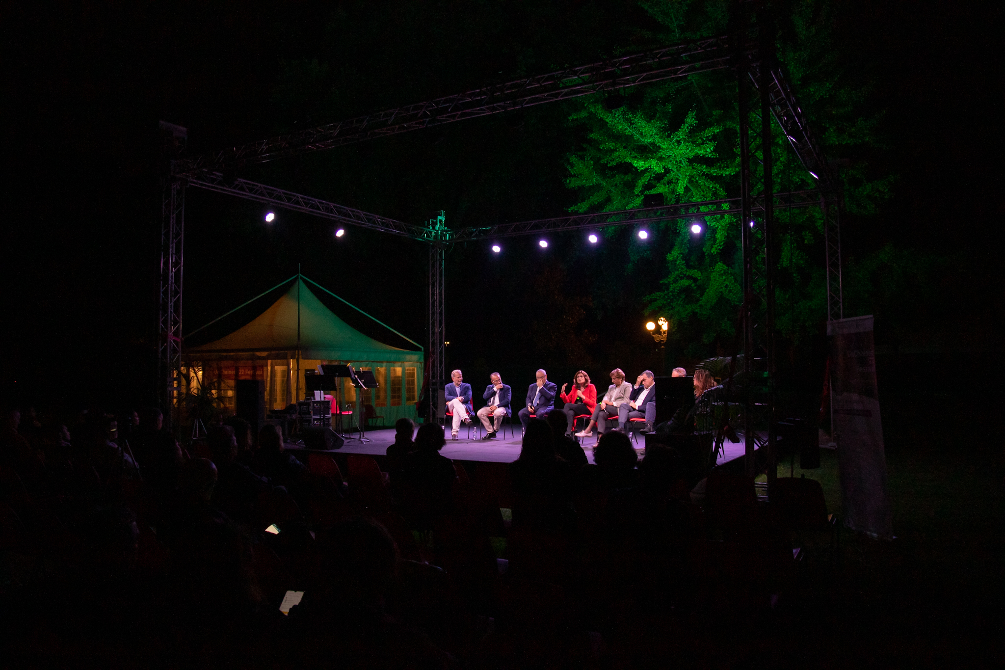 Discussione notturna su palco all'aperto con relatori illuminati da luci sceniche e tenda luminosa sullo sfondo.