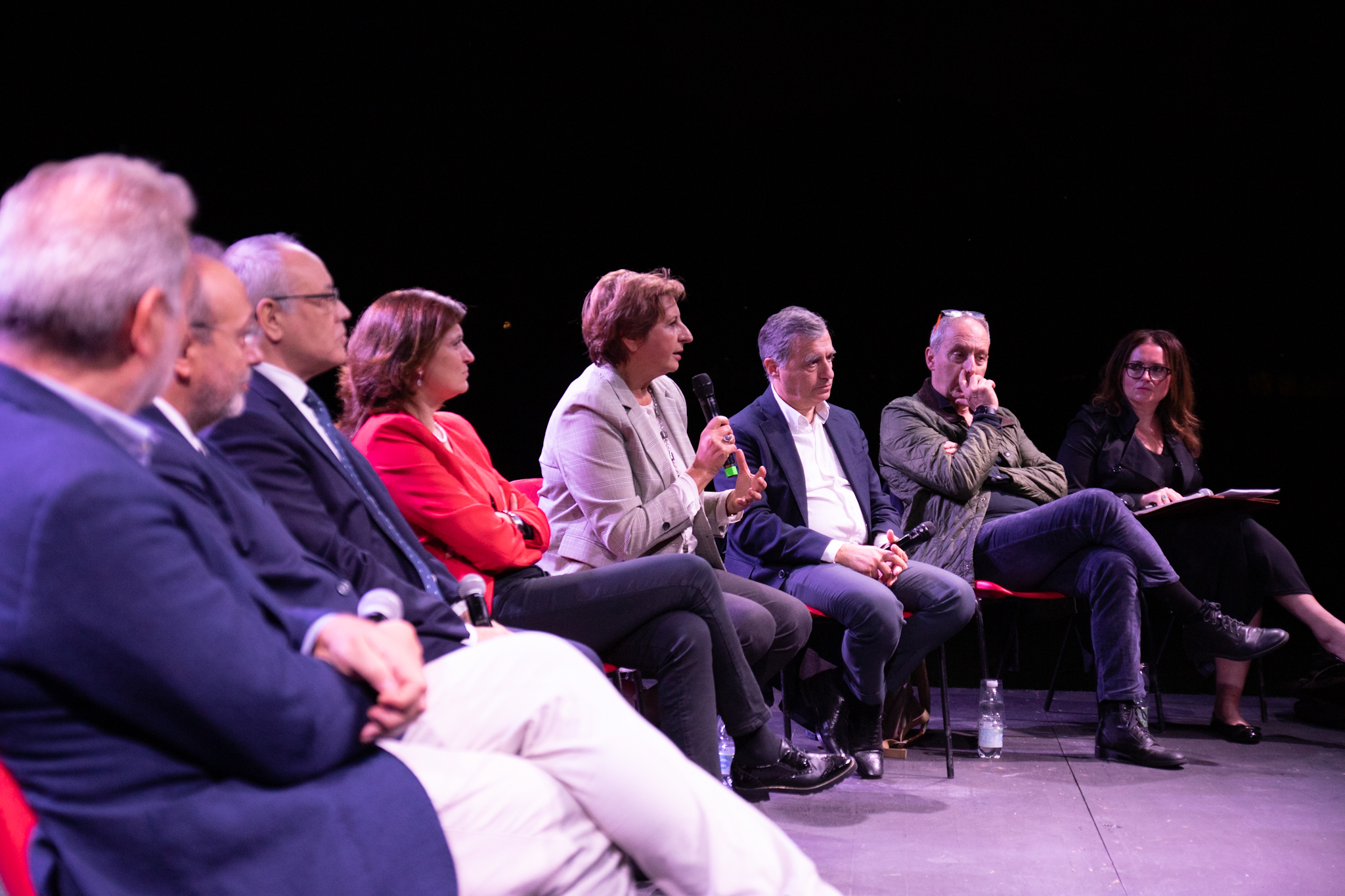 Gruppo di persone sedute su un palco durante una discussione pubblica, una donna parla con un microfono in mano.