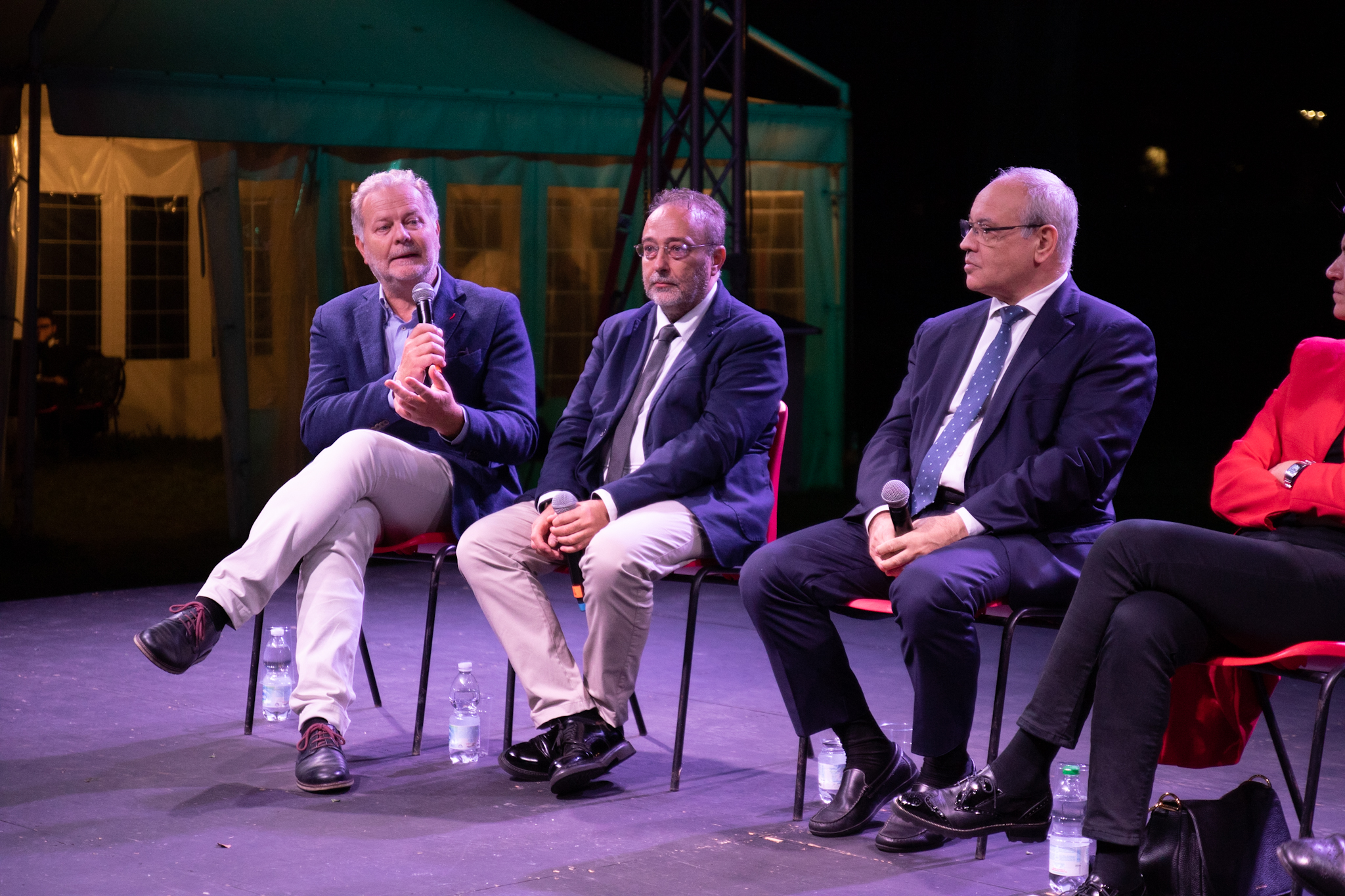 Quattro persone sedute su un palco, partecipano a una discussione pubblica serale, microfoni in mano.