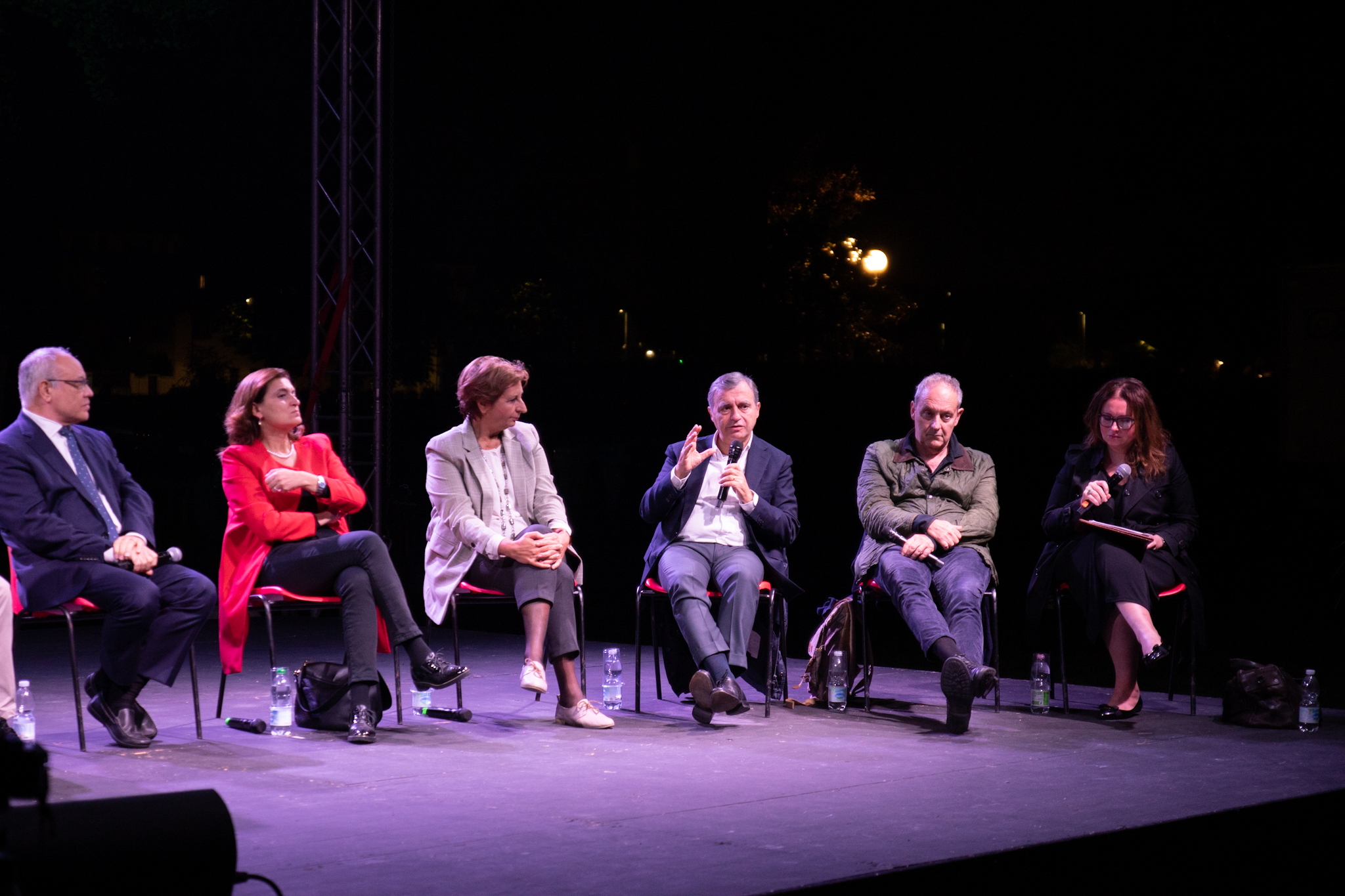 Gruppo di persone sedute su un palco durante una discussione pubblica serale, con luci sullo sfondo.