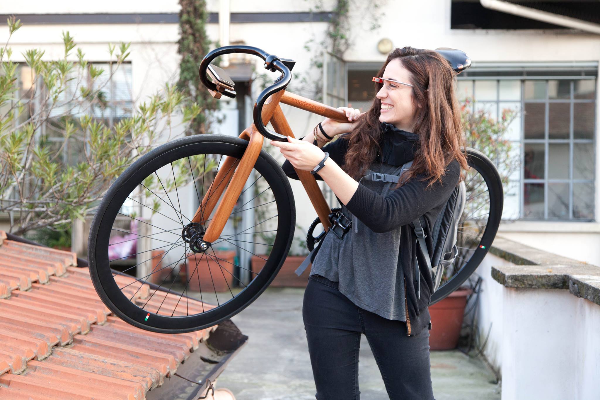 Donna sorridente con occhiali tecnologici, porta una bicicletta di legno alzata su una spalla, sfondo urbano.