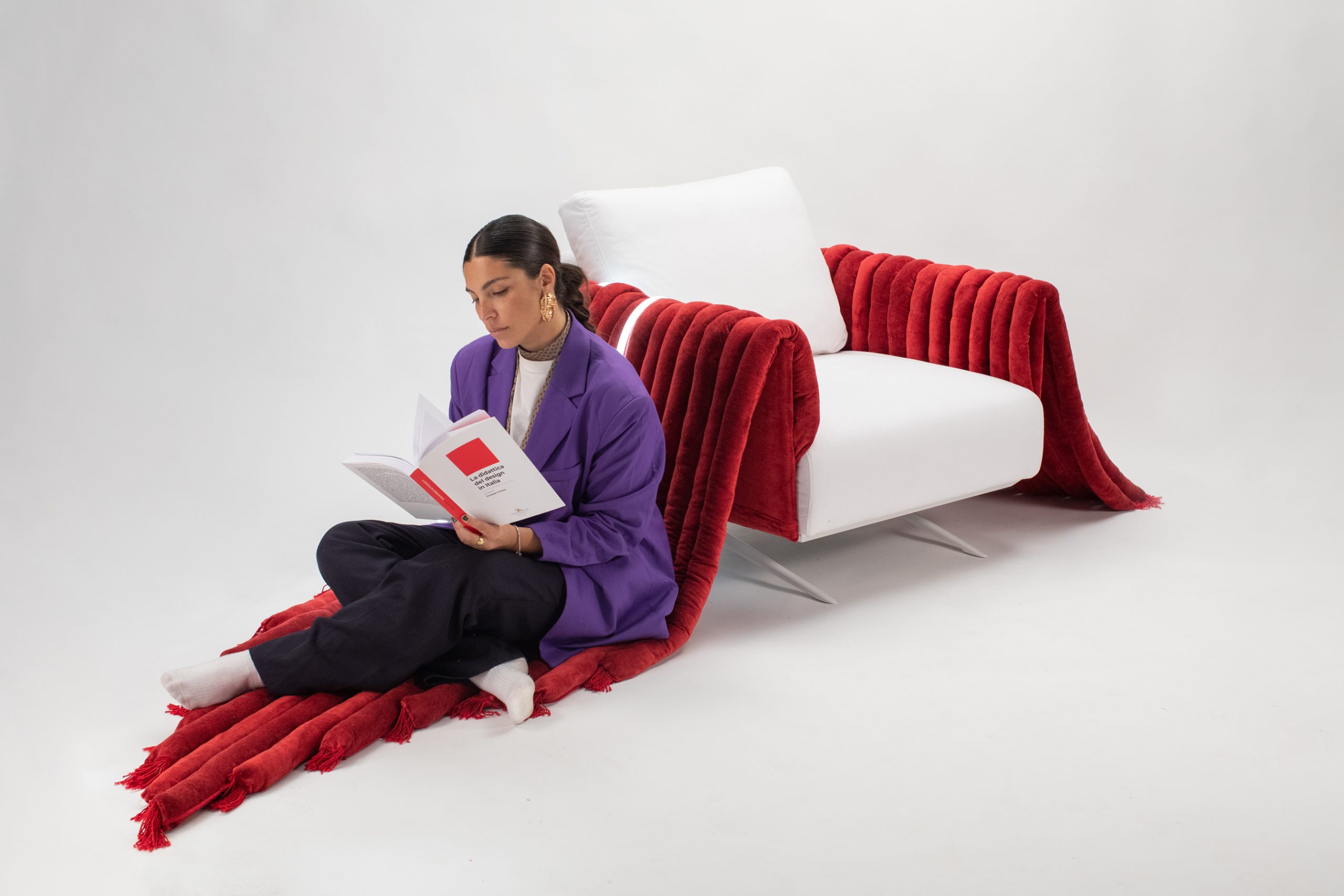 Persona seduta su tappeto rosso, legge libro accanto a poltrona bianca con coperta rossa. Ambiente minimalista.