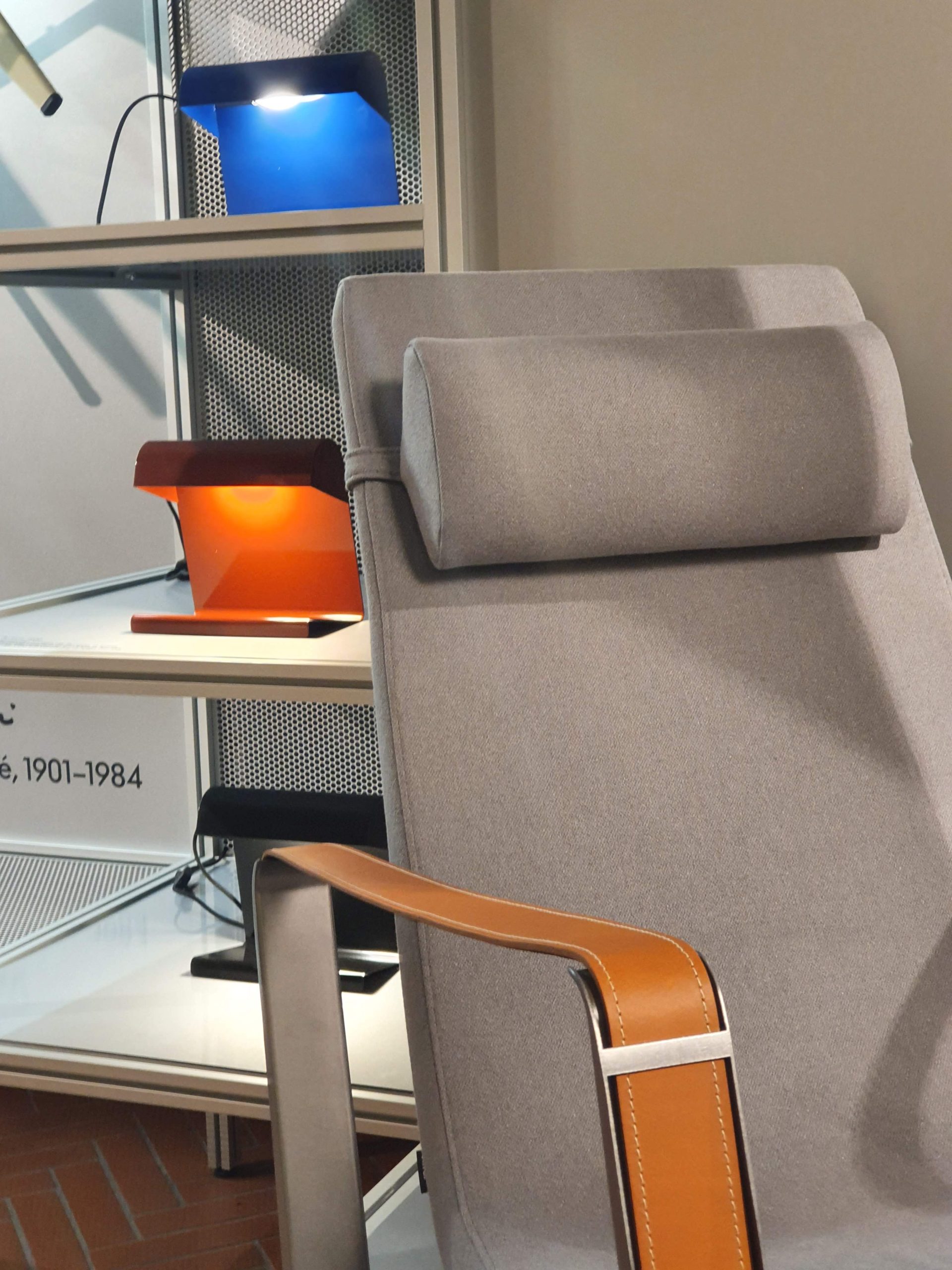 Poltrona moderna in tessuto grigio con bracciolo in cuoio, accanto a lampade di design blu e arancioni su mensole.