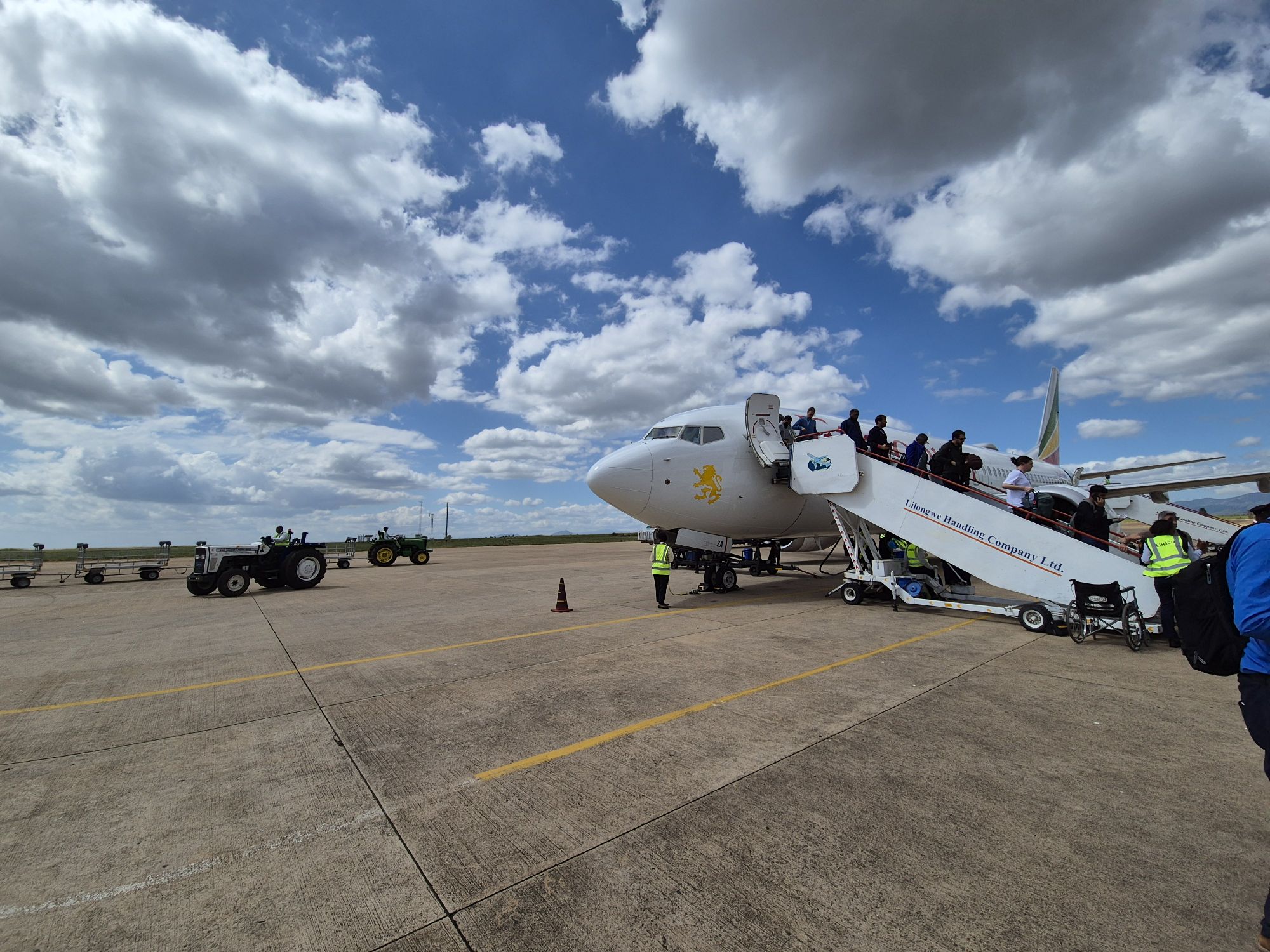 Passeggeri salgono a bordo di un aereo bianco su pista aeroportuale sotto un cielo nuvoloso.