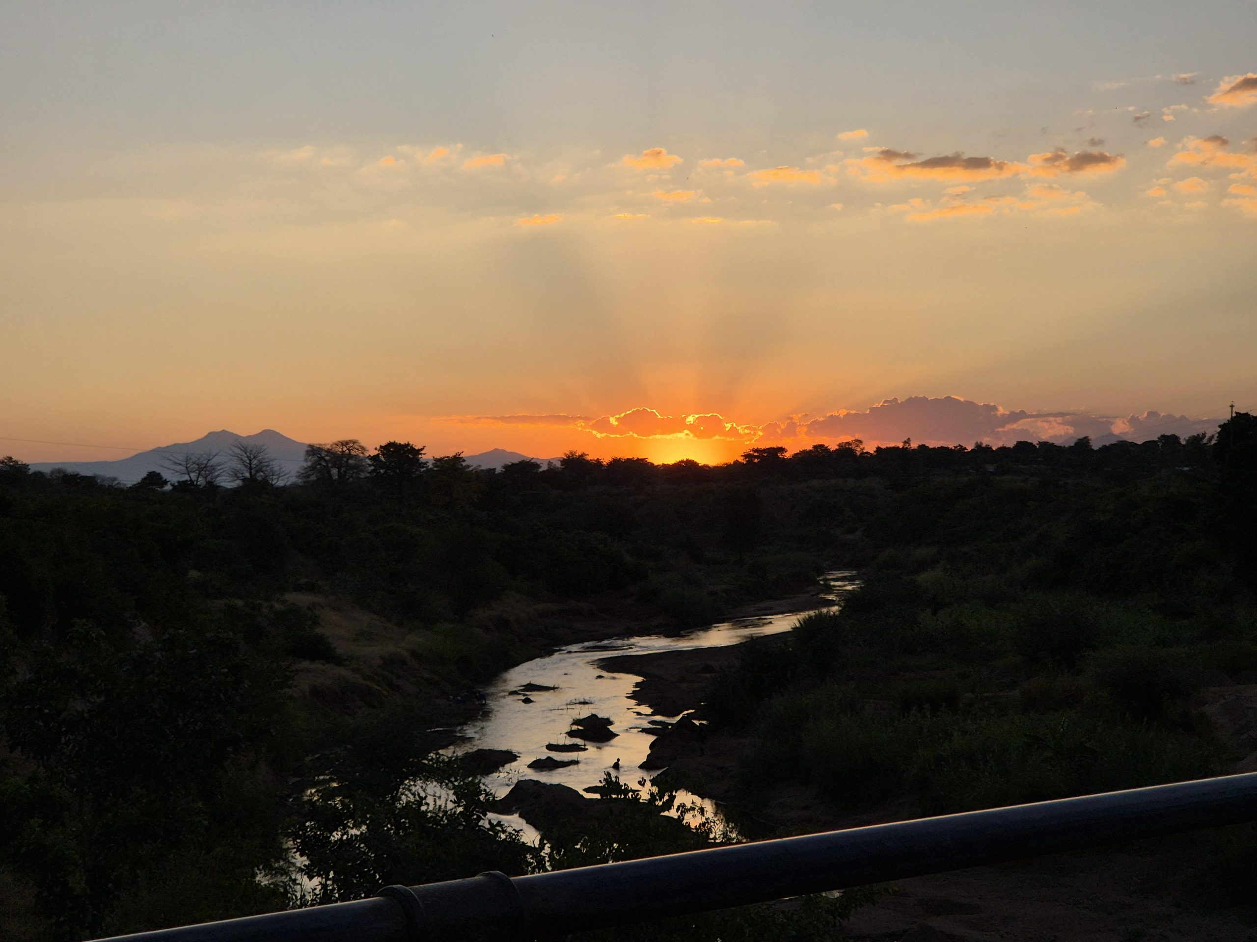 Tramonto su un fiume serpeggiante, con montagne all'orizzonte e cielo arancione, nuvole e silhouette degli alberi.