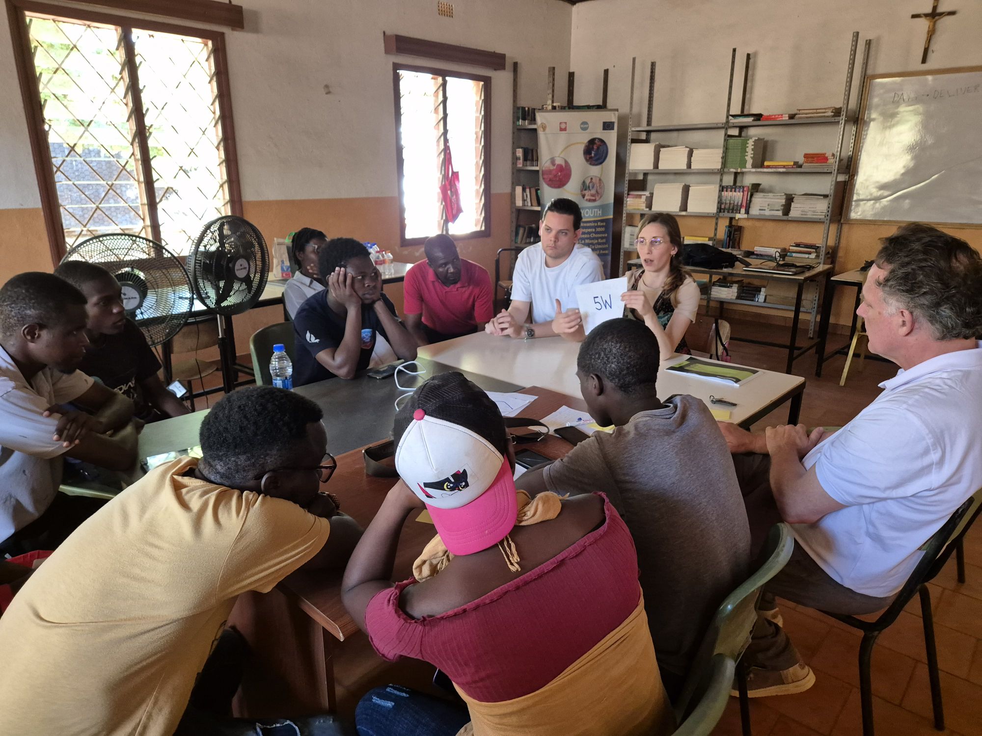 Gruppo di persone in riunione in aula luminosa, discutendo attorno a un tavolo con libri e lavagna sullo sfondo.