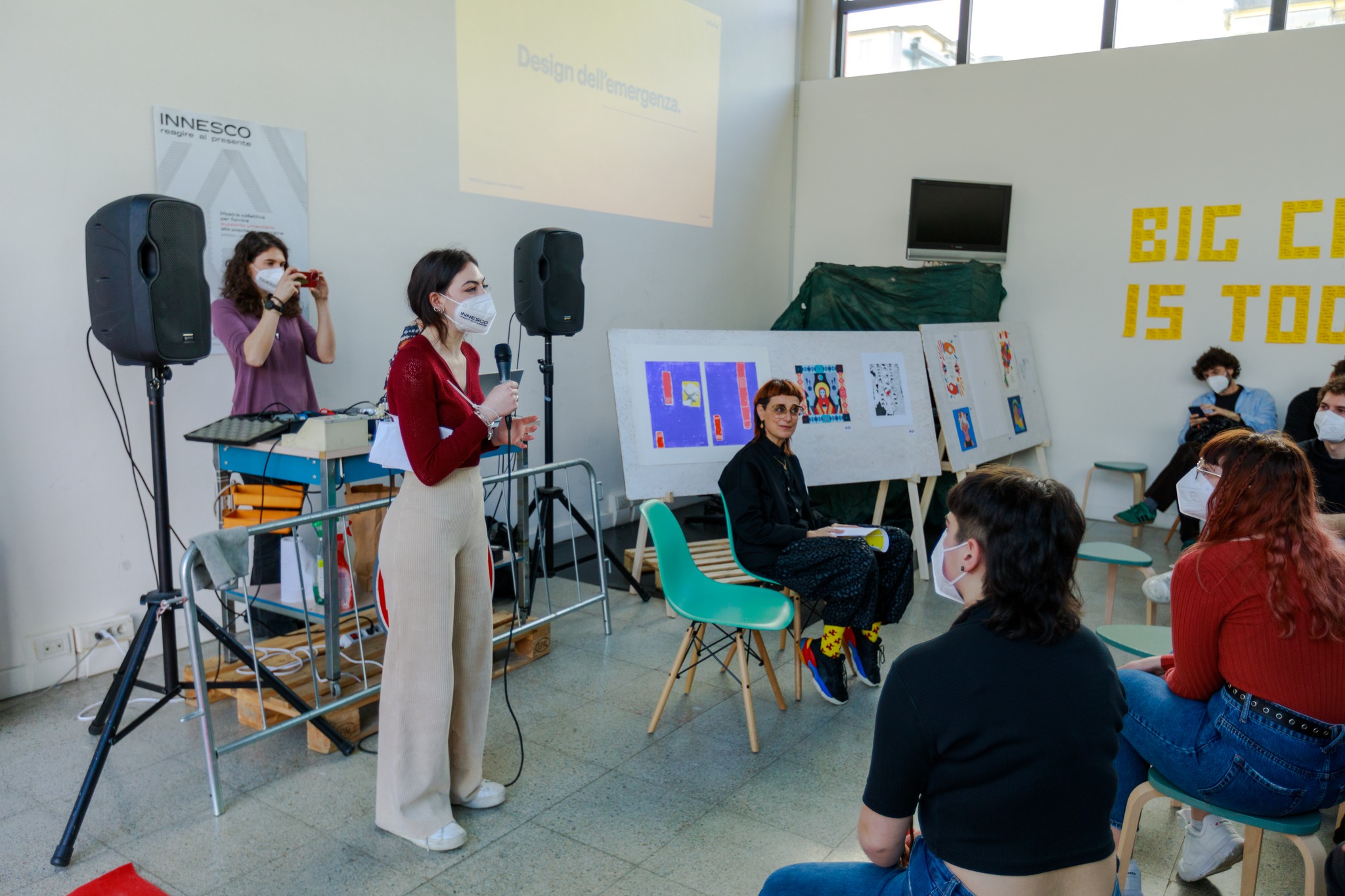 Persone partecipano a un workshop di design in una sala, con opere d'arte esposte sullo sfondo.