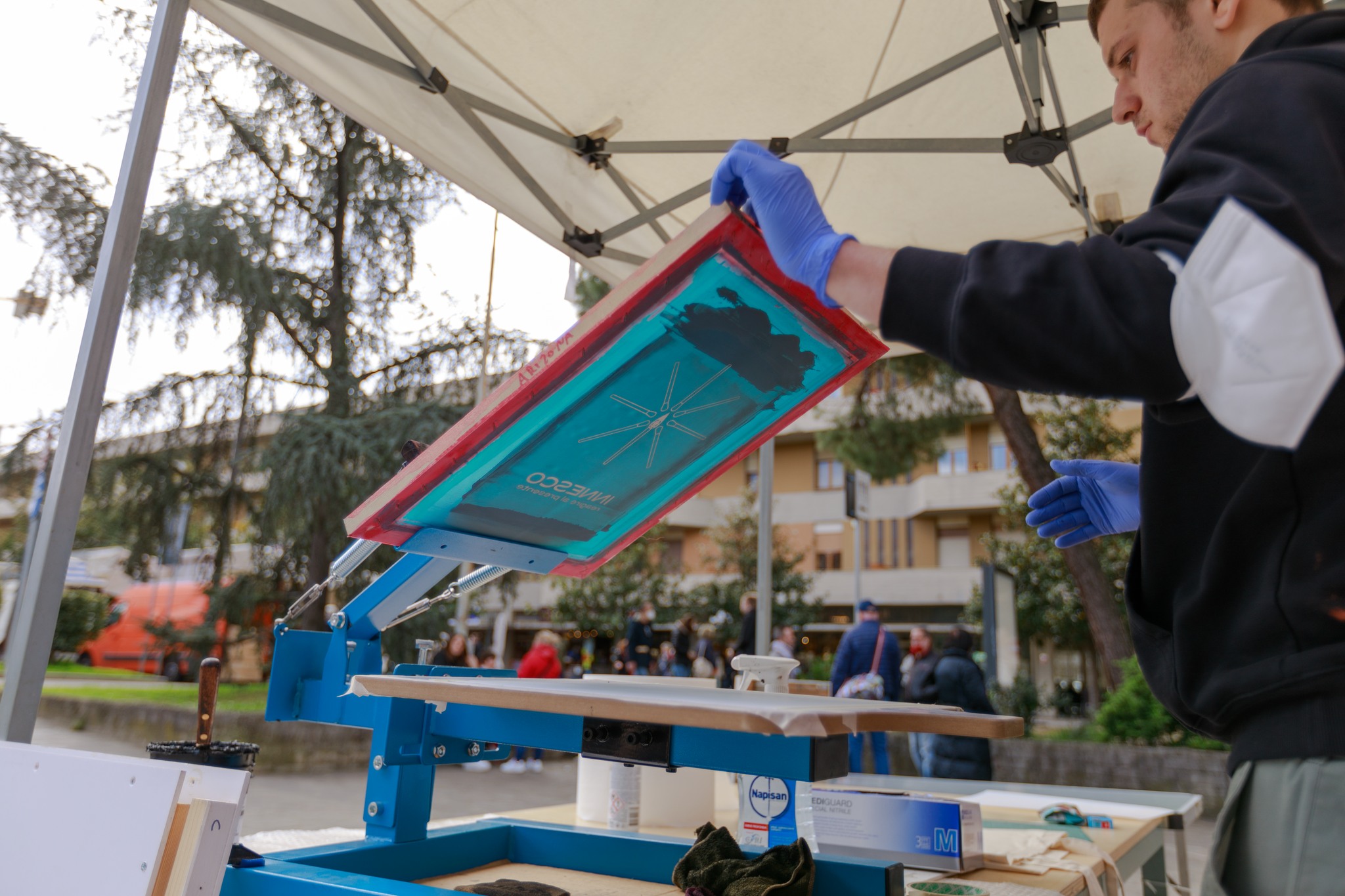 Persona all'aperto prepara scena di serigrafia sotto tendone, sfondo urbano e naturale.