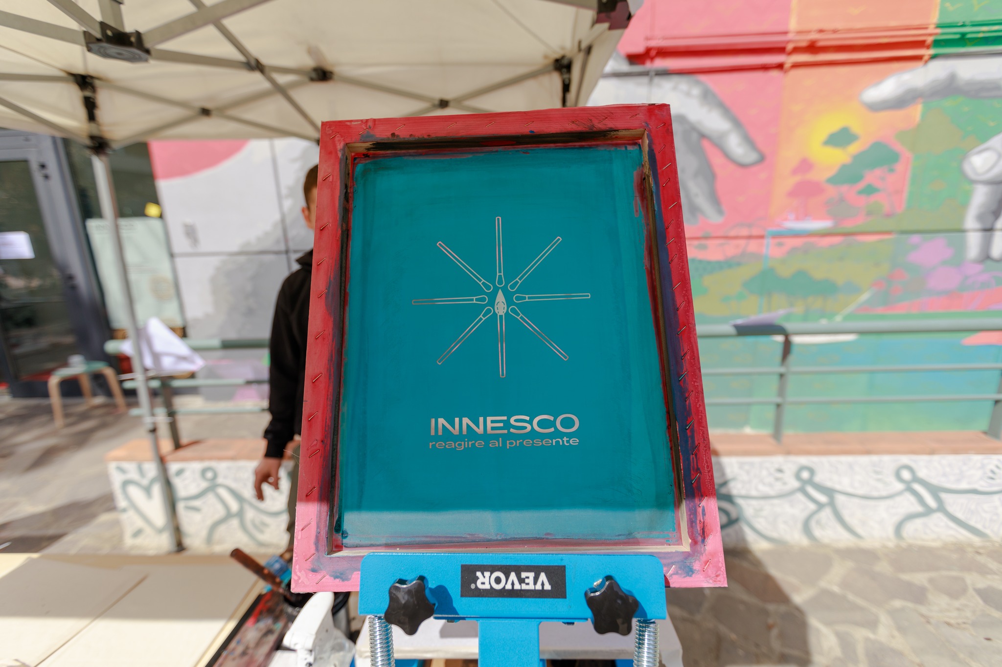 Schermo per serigrafia con logo INNESCO e sfondo colorato di un murale artistico all'aperto.