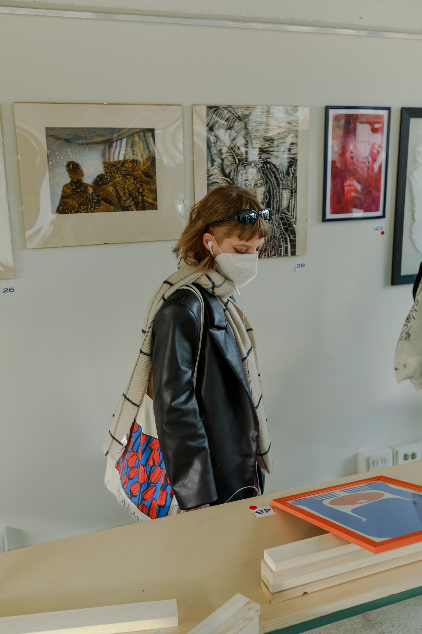 Persona con mascherina e occhiali da sole osserva opere d'arte in una galleria, indossa giacca in pelle e sciarpa.