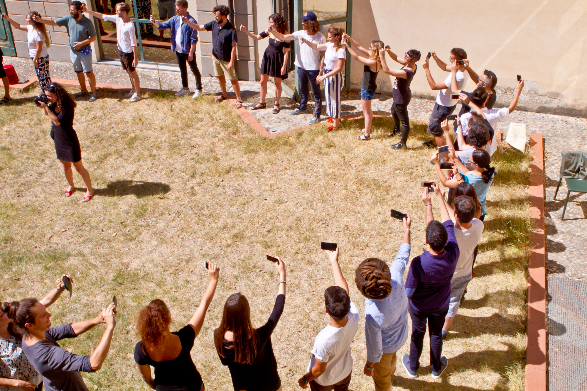 Persone in cerchio con smartphone scattano foto, una donna al centro con una fotocamera su prato.