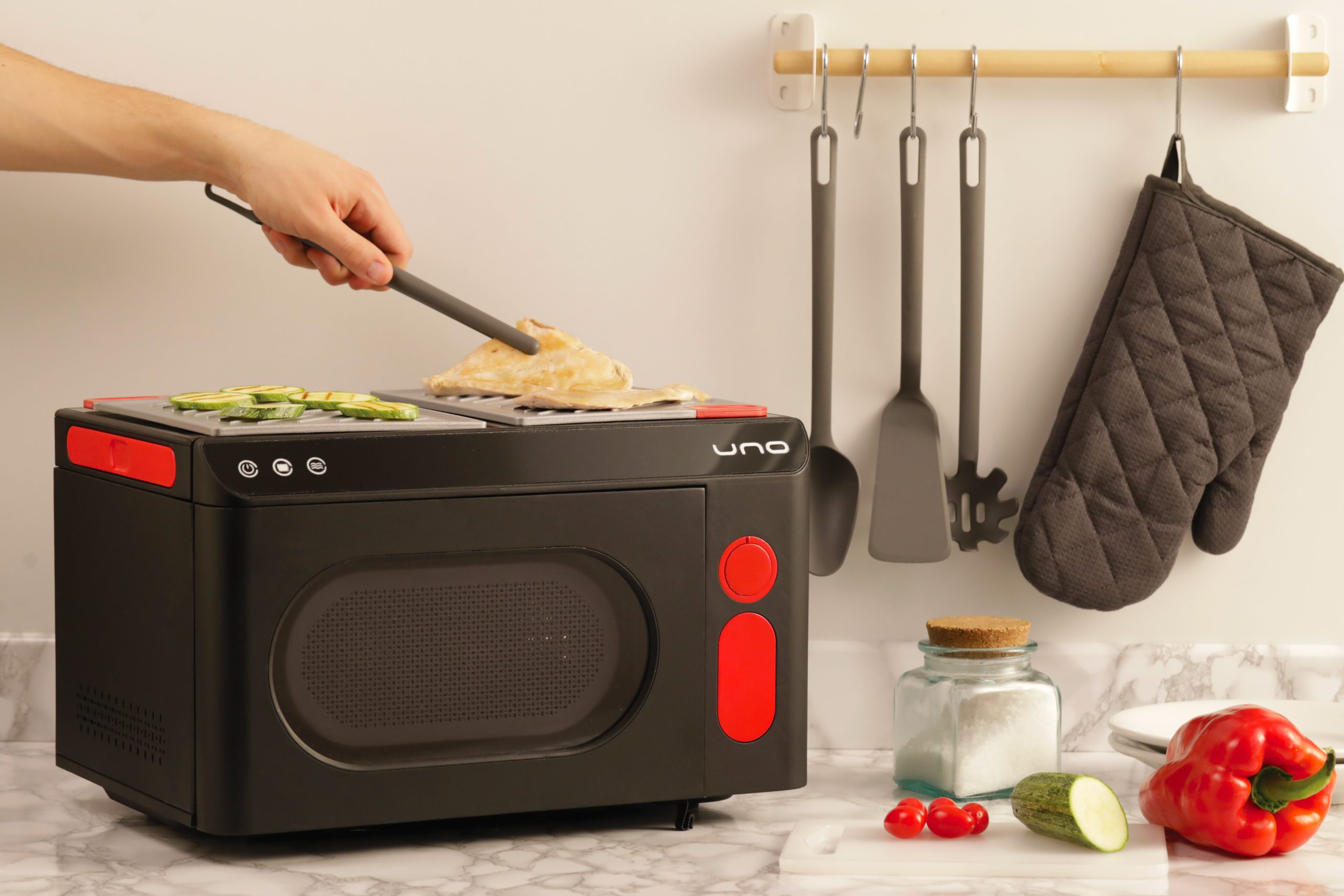 Persona cucina pollo su un piccolo forno nero con verdure, utensili appesi e ingredienti sulla superficie di marmo.