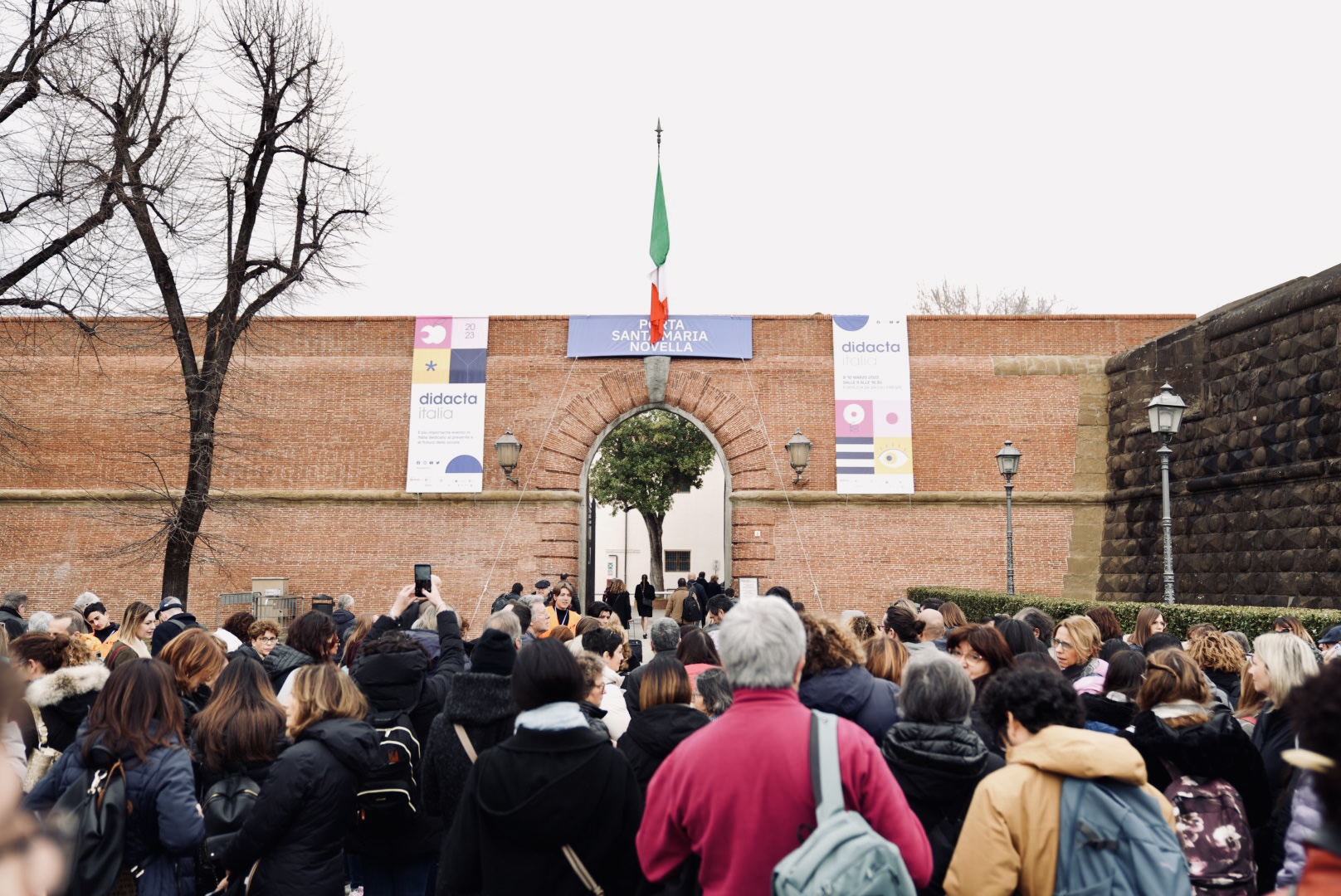 Ingresso fiera Didacta Italia 2023 con folla di visitatori sotto bandiera italiana a Fortezza da Basso, Firenze.