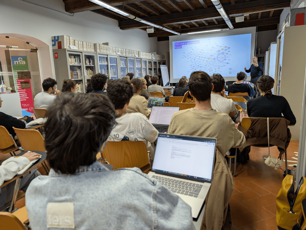 Persone in aula assistono a una presentazione con diagramma su schermo, con laptop aperti su scrivanie.