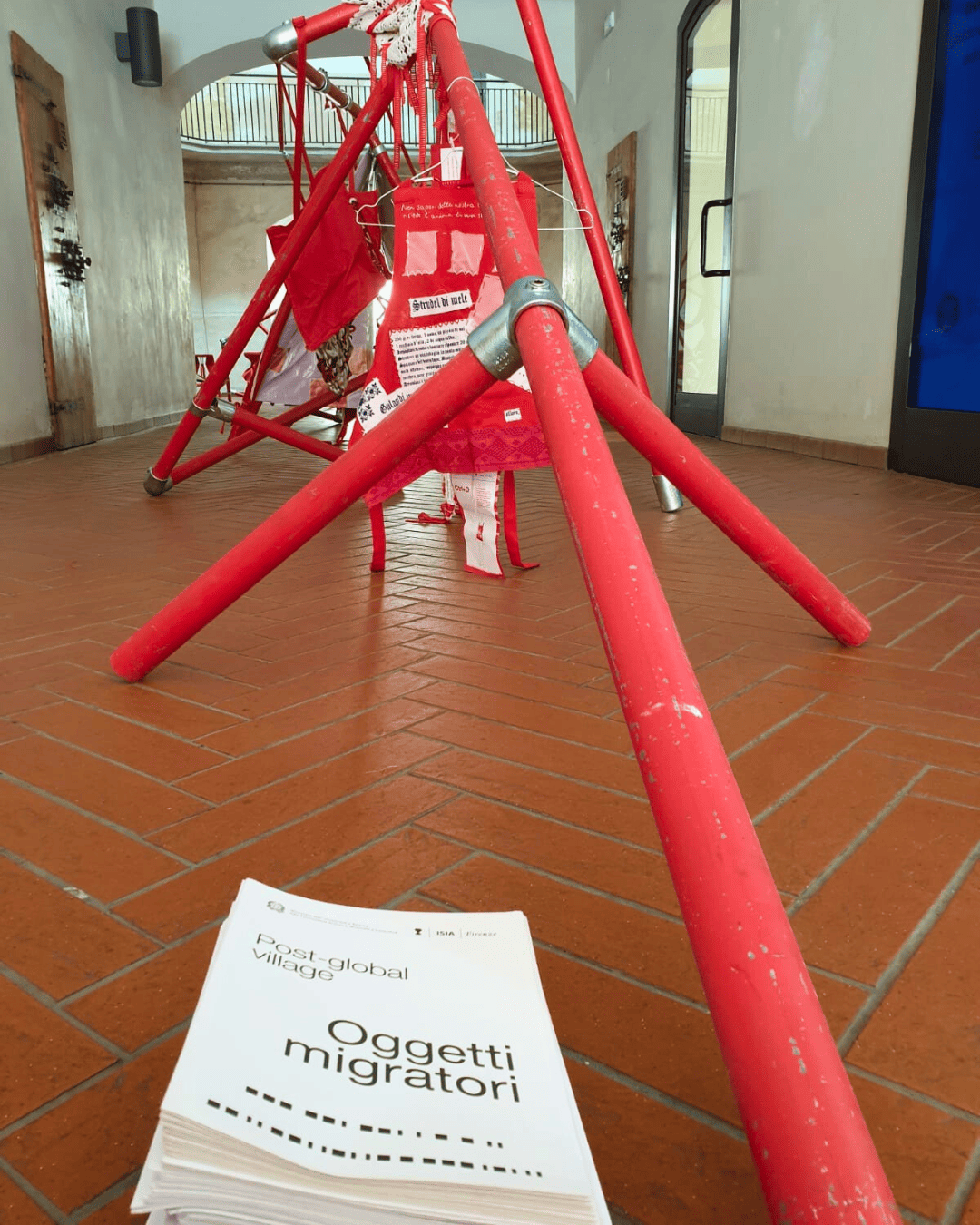 Esposizione d'arte contemporanea con installazione rossa e brochure intitolata Oggetti migratori su pavimento in cotto.