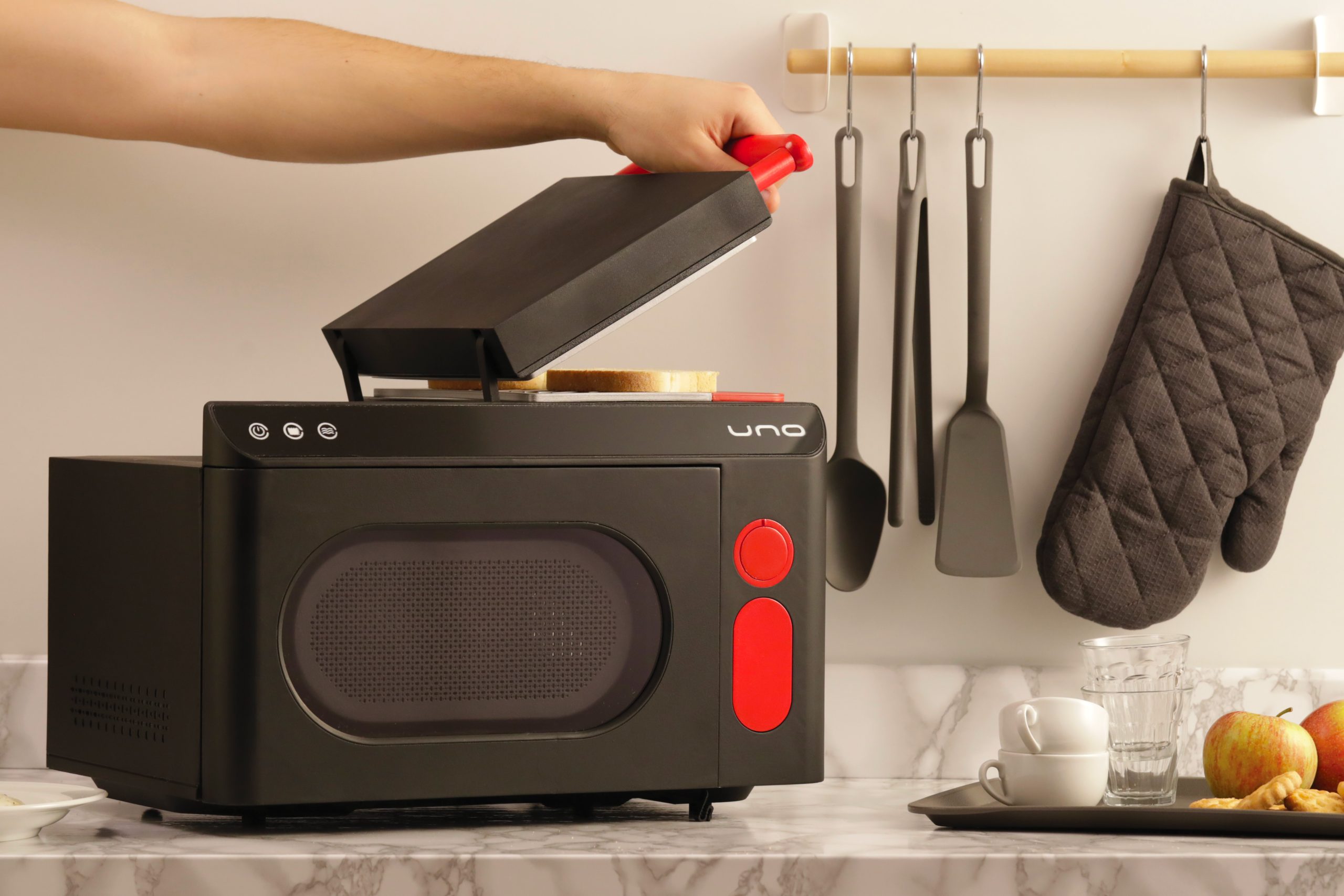 Tostapane nero su piano cucina in marmo con utensili appesi e tazze a lato, un toast è in preparazione.