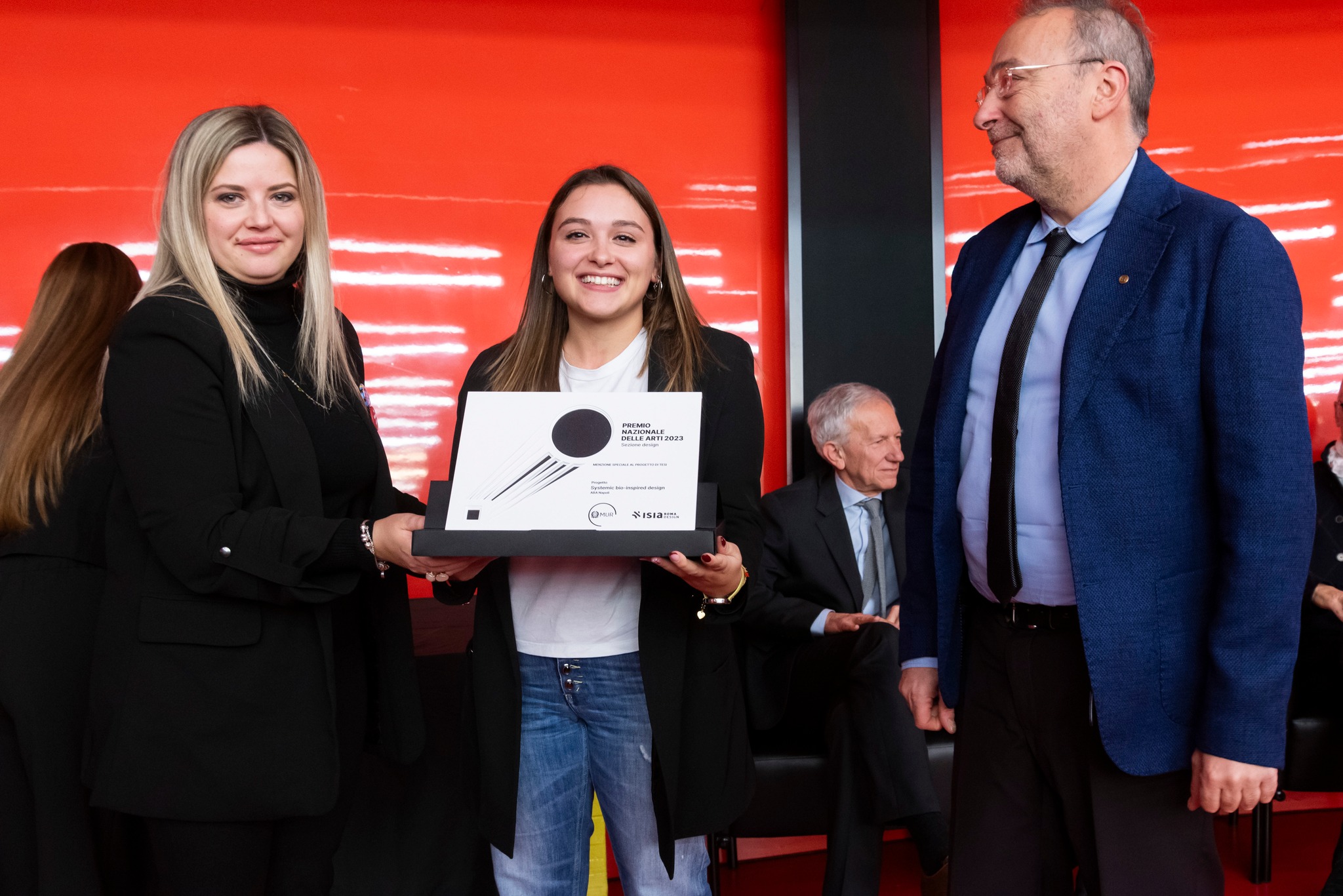 Donna riceve un premio su sfondo rosso mentre altre persone sorridono alla cerimonia.
