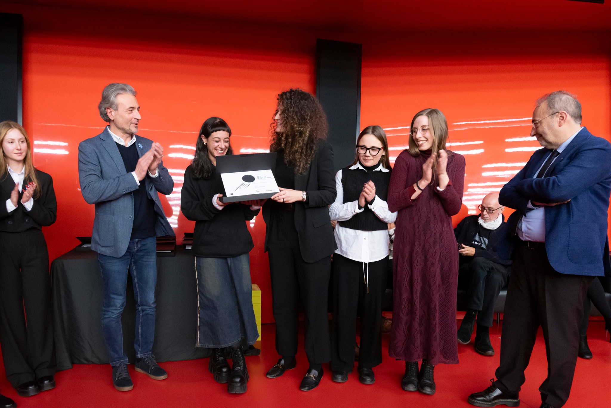 Premiazione con persone sorridenti su sfondo rosso, mentre viene consegnato un riconoscimento.