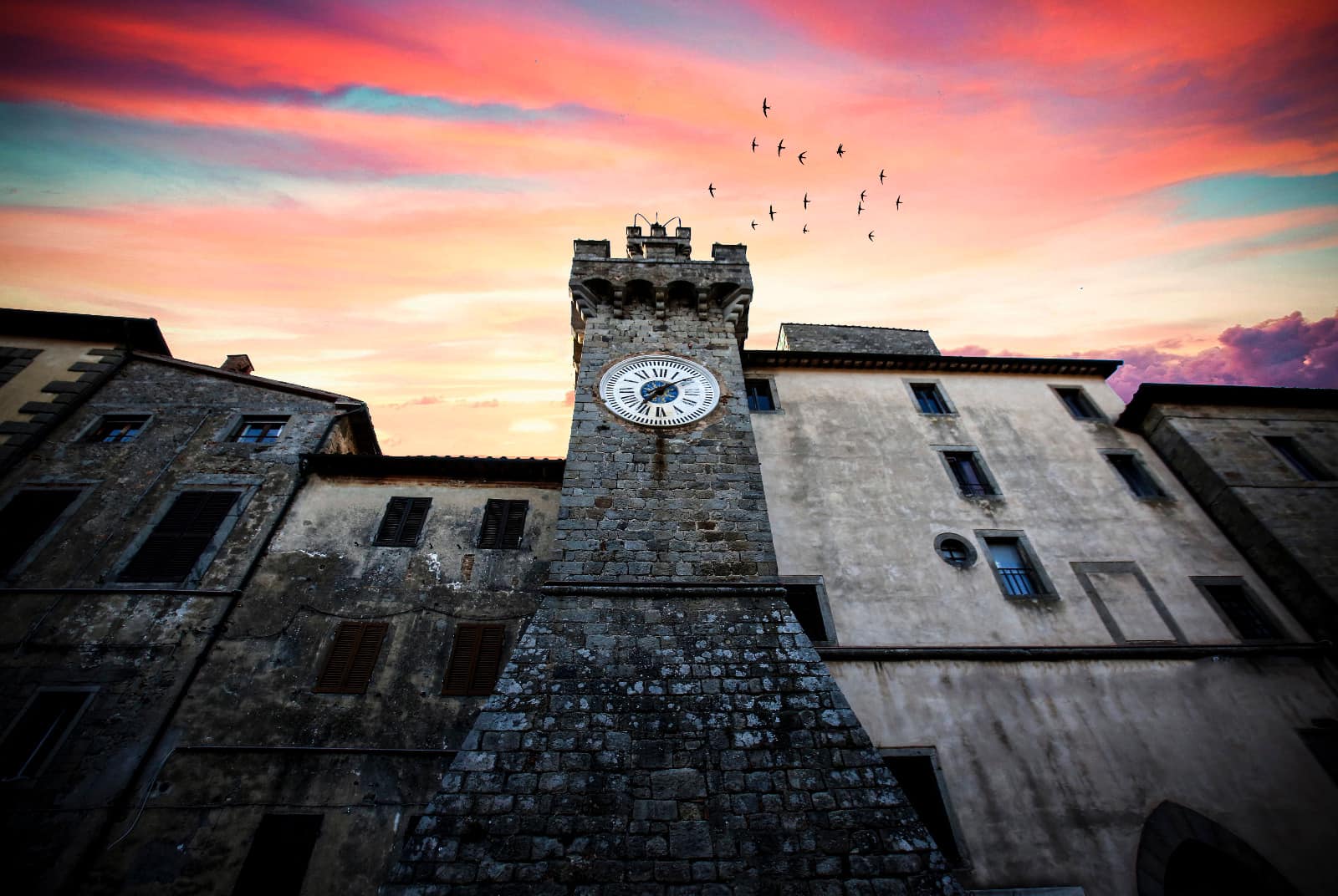 Torre medievale con orologio al tramonto, cielo colorato e stormo di uccelli in volo.