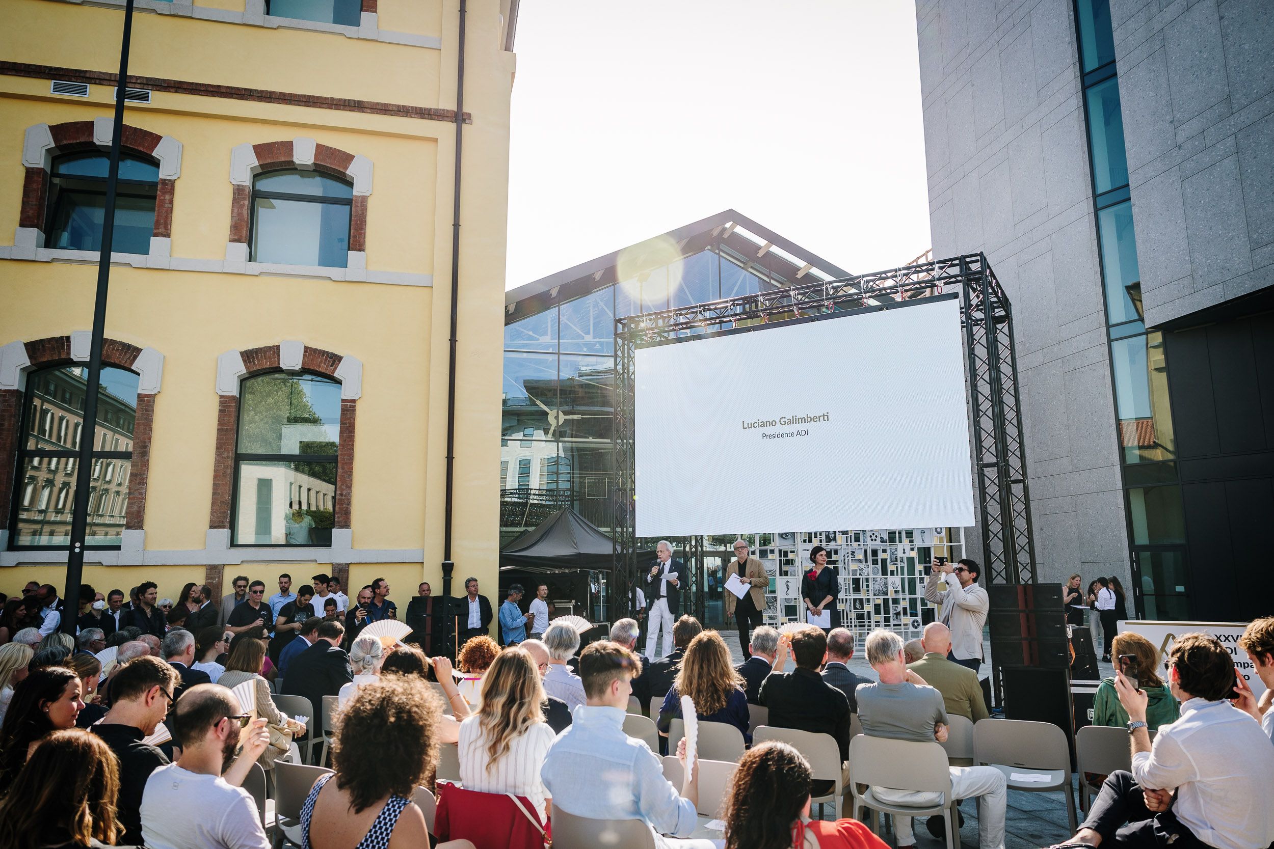 Evento all'aperto con folla e maxischermo, vicino a edifici moderni. Luci solari illuminano la scena.
