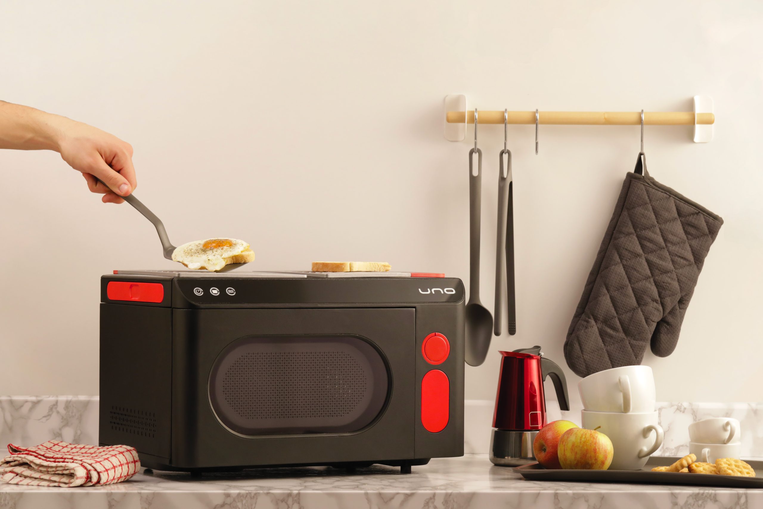 Mano prepara uovo fritto su cucina compatta, con utensili appesi. Tazze e mele su bancone marmo.