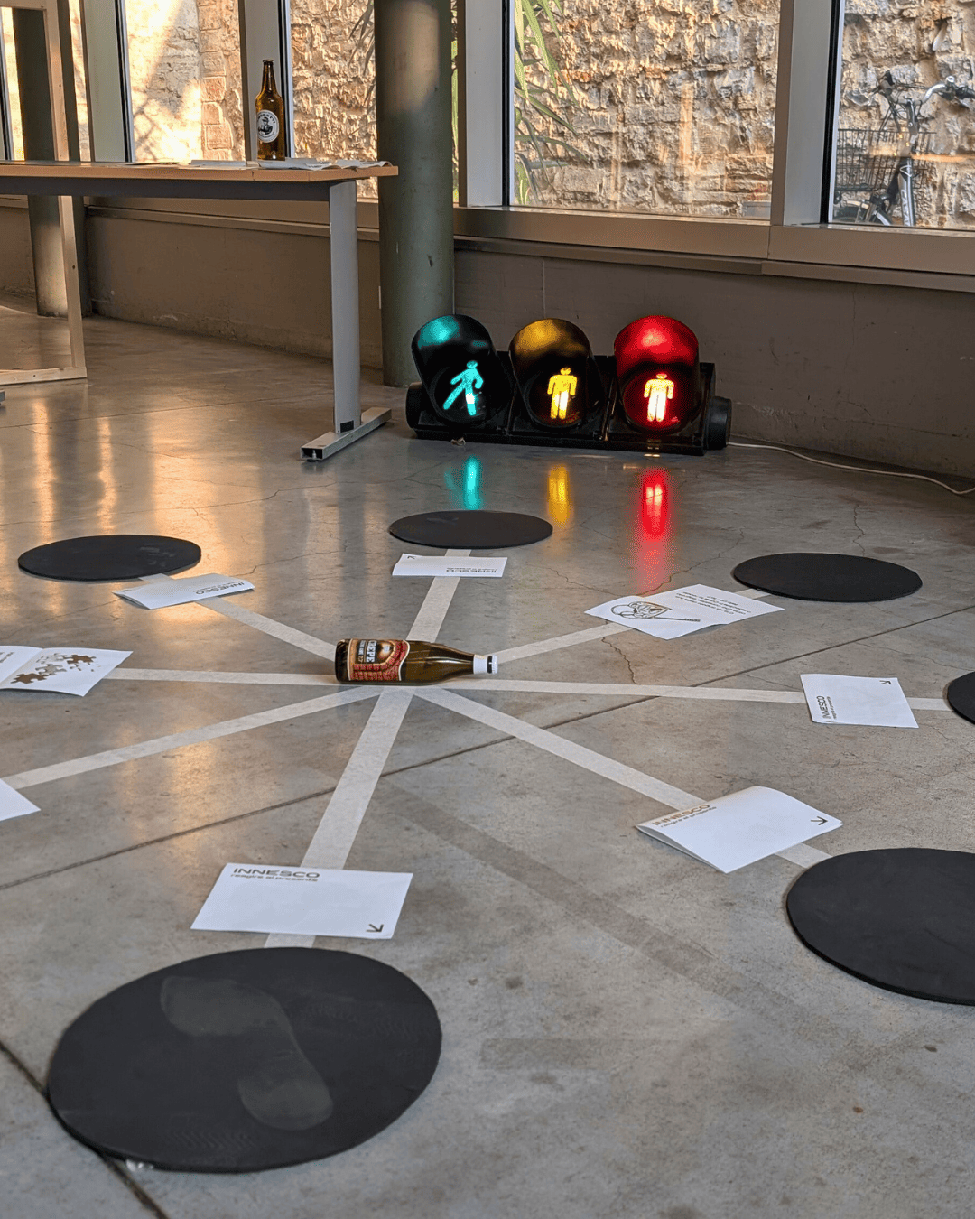 Gioco della bottiglia con birra al centro, semaforo e fogli su pavimento in cemento di uno spazio industriale.