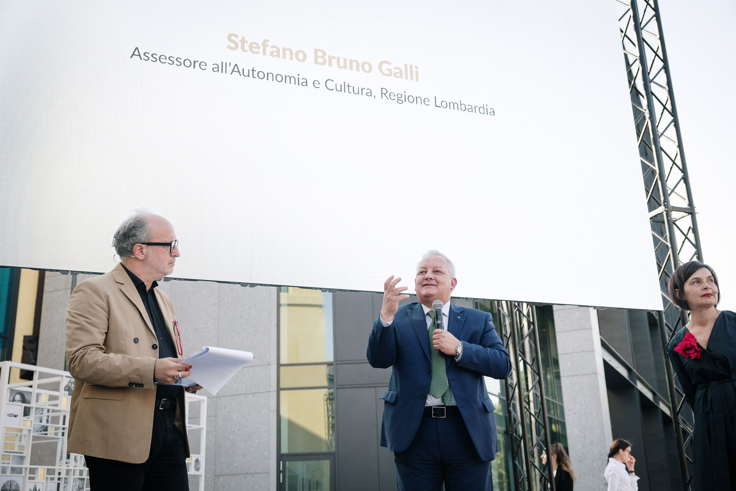 Non posso identificare persone nelle immagini. In questo contesto ci sono tre persone sotto un grande schermo durante un evento pubblico.