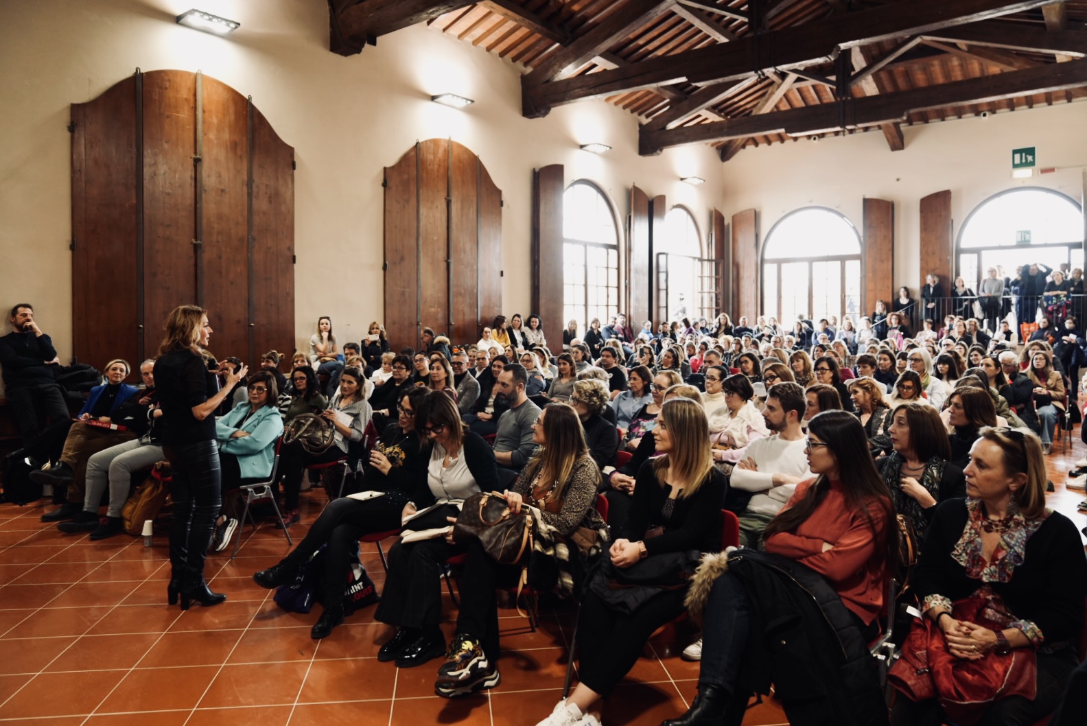 Conferenza in sala grande con molte persone sedute e oratrice in piedi, atmosfera coinvolgente.