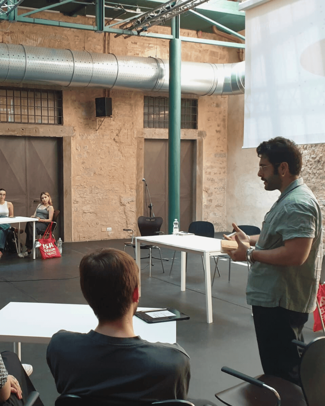 Una persona parla a un gruppo di studenti seduti attorno a tavoli in una sala con pareti in mattoni e tubi a vista.