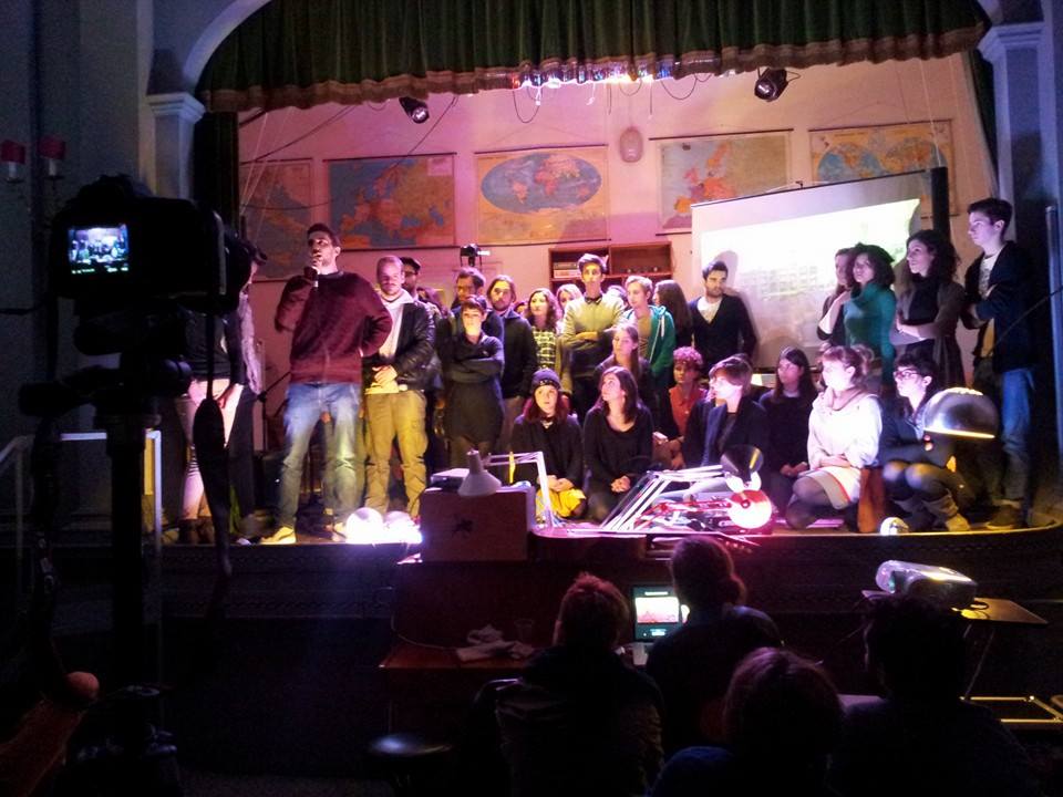 Gruppo di persone su palco per presentazione, con mappe geografiche sullo sfondo e luci di scena calde.