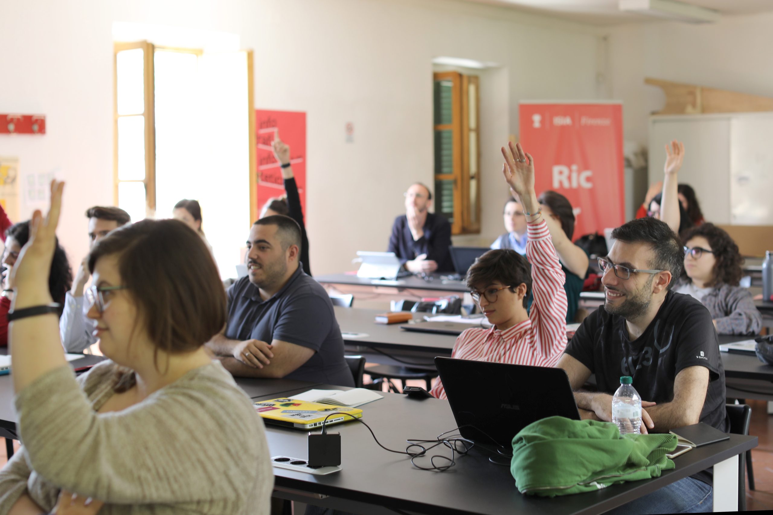 Studenti in aula con laptop, alcuni alzano la mano durante una lezione interattiva, ambiente luminoso ed educativo.