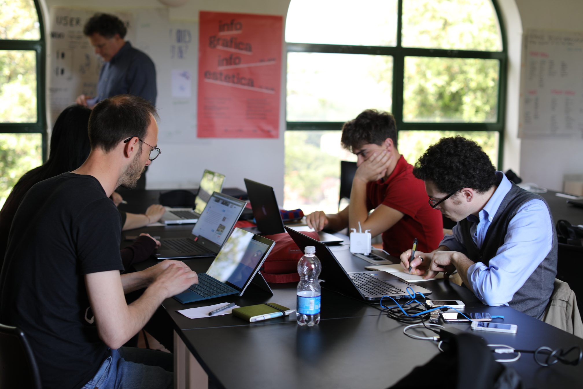 Persone al lavoro su laptop in un'aula luminosa con poster alle pareti, concentrati su compiti creativi e studio.