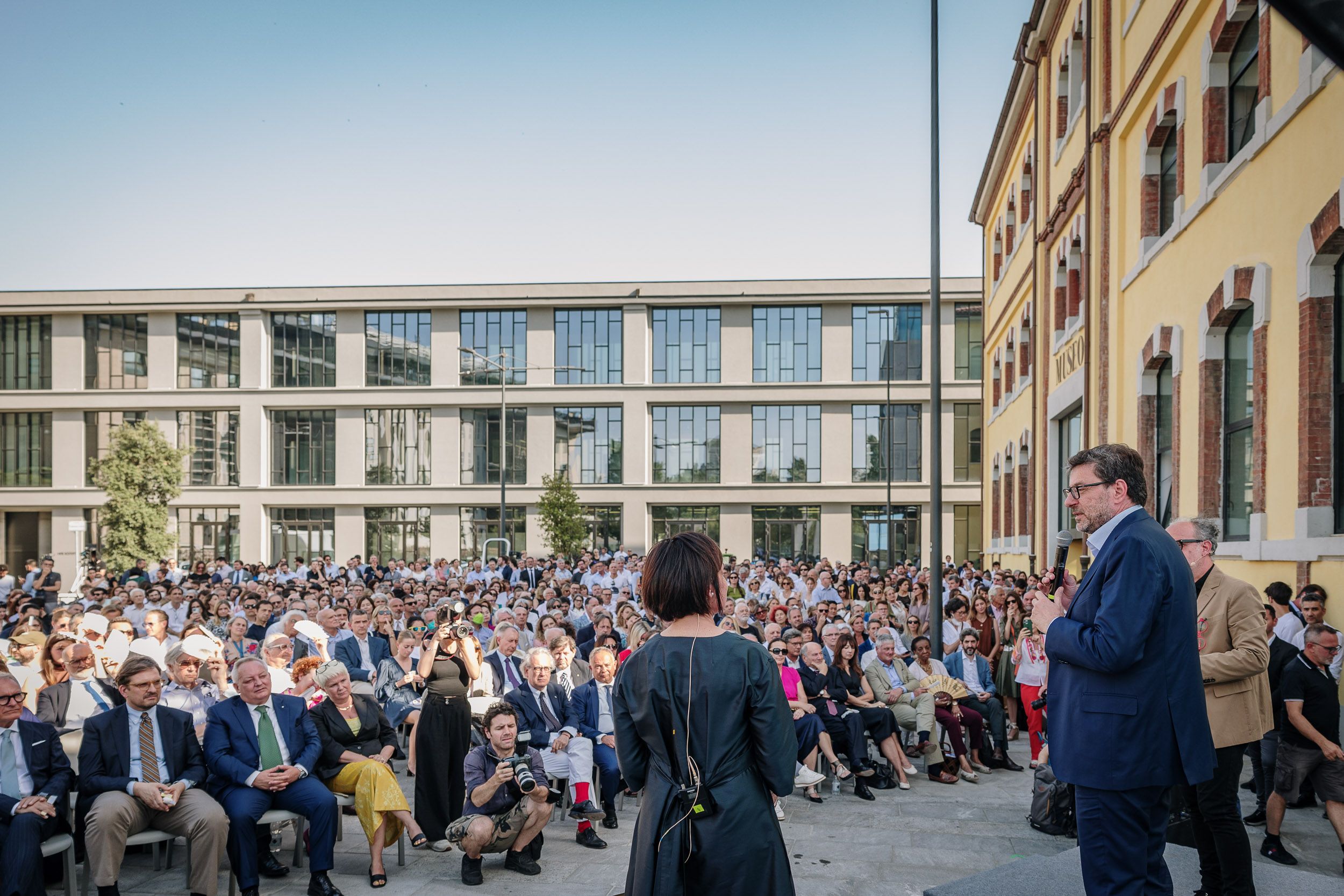 Assemblea pubblica all'aperto con oratori sul palco e numerosi partecipanti seduti davanti a un edificio moderno.