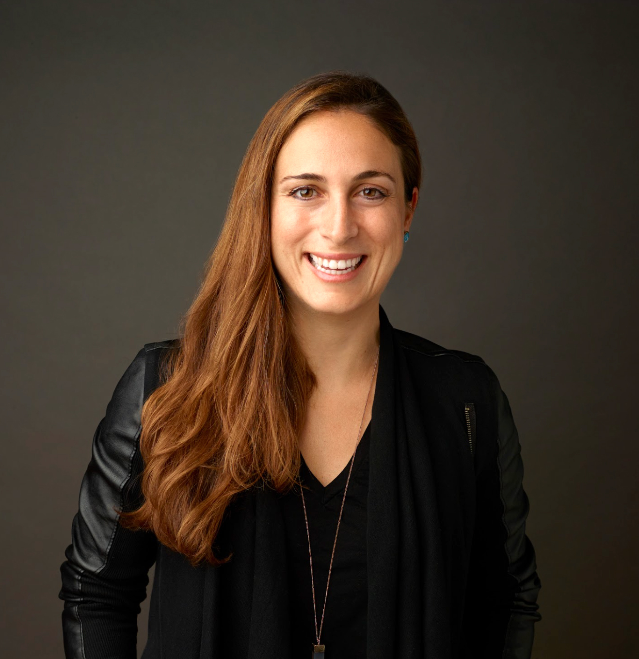 Donna sorridente con capelli lunghi e castani, indossa giacca nera, fondo nero neutro.