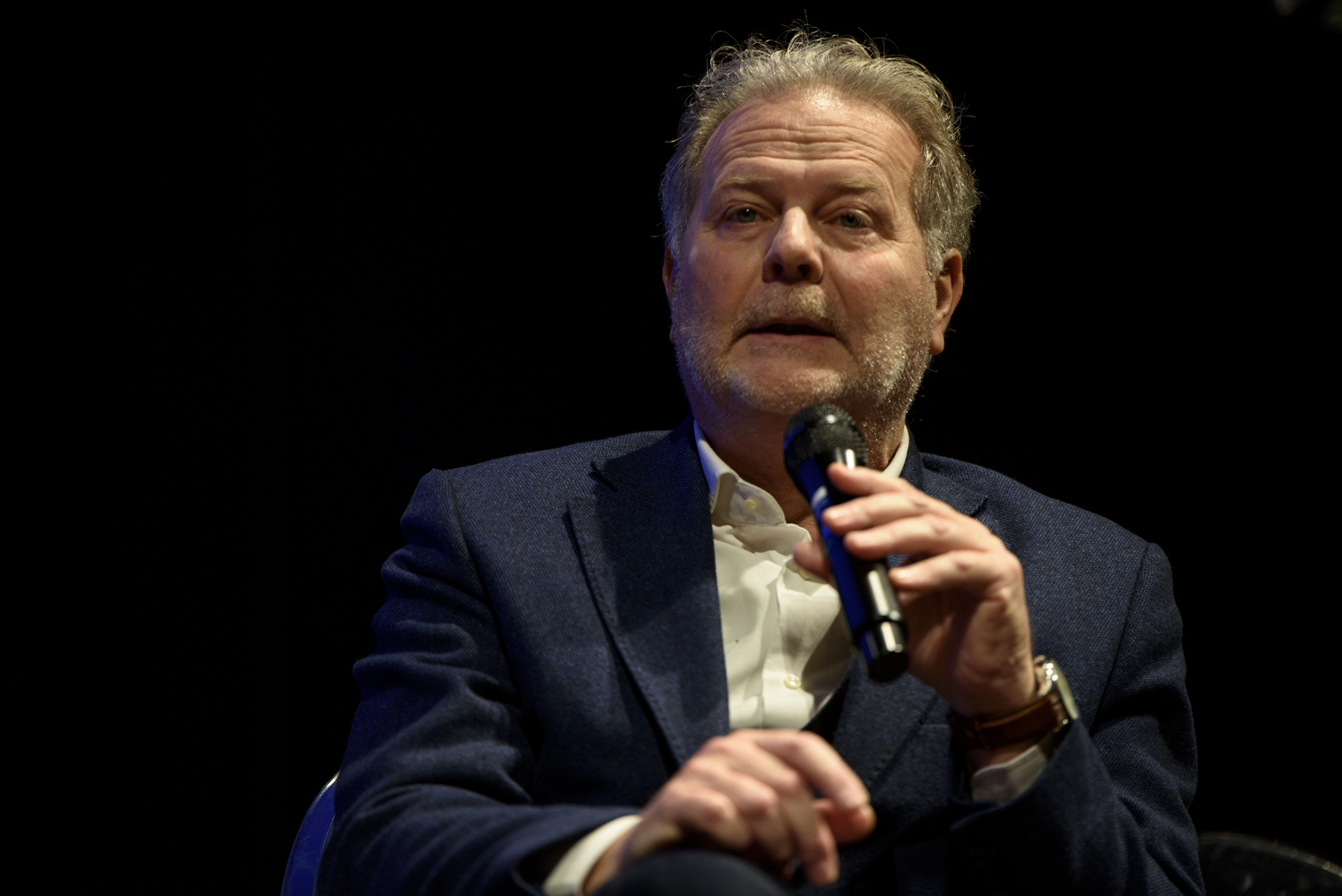 Uomo con microfono, in giacca blu e camicia bianca, parla su sfondo nero.