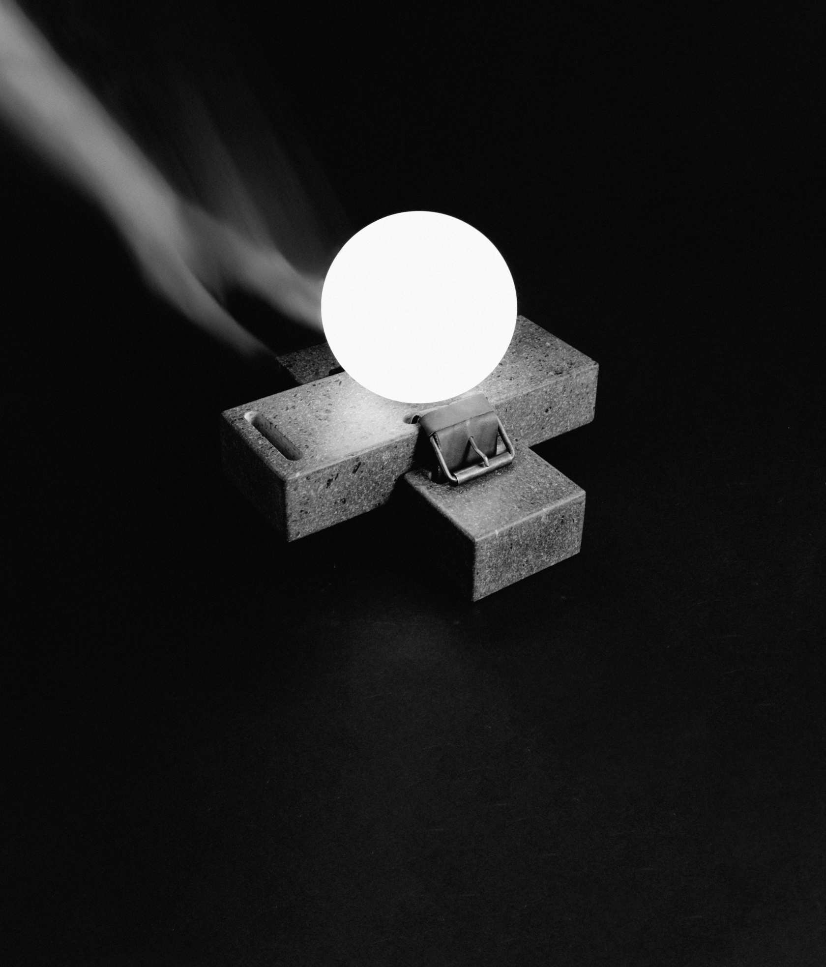 Lampada a sfera luminosa su base in cemento a forma di croce, in bianco e nero su sfondo scuro.