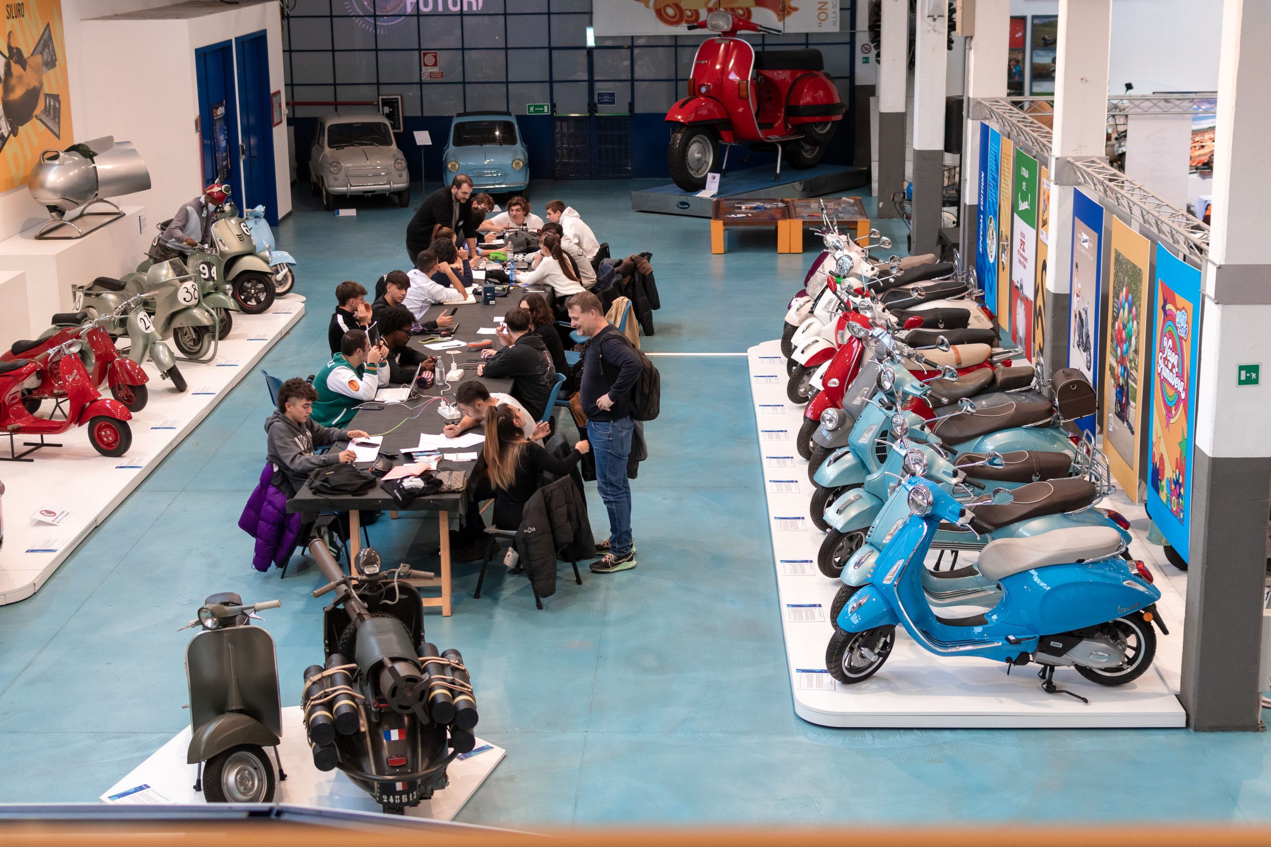 Persone in riunione tra file di scooter e Vespe in un museo, atmosfera di lavoro e esposizione storica.