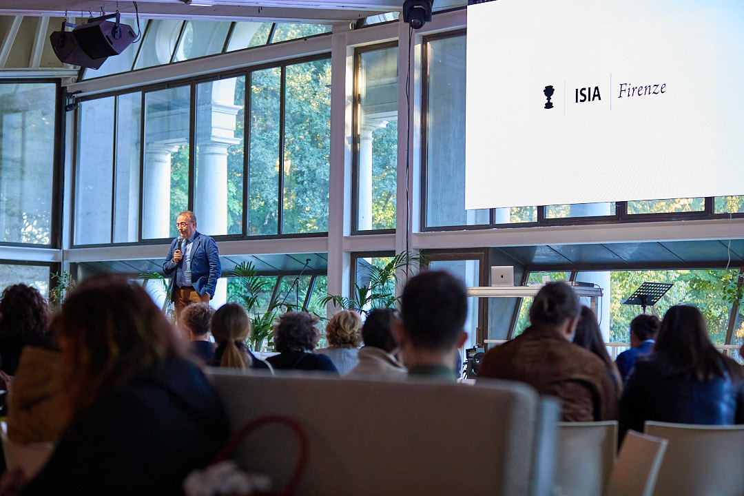 Uomo su palco parla a un pubblico in sala conferenze con vetrate e schermo proiettante logo ISIA Firenze.