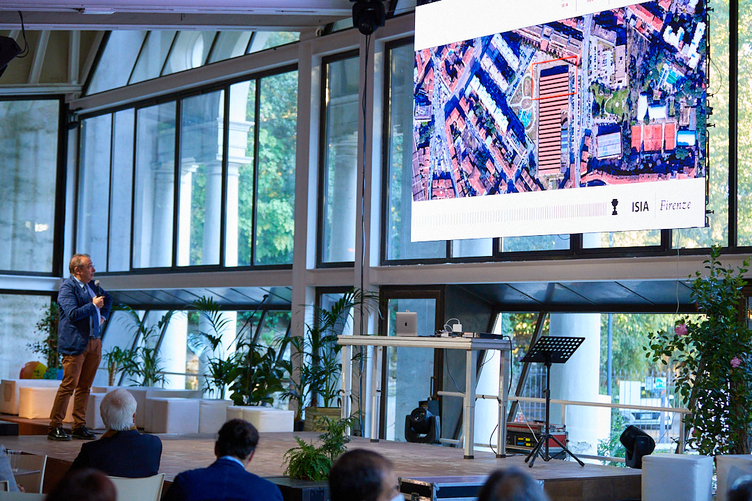 Oratore presenta una mappa satellitare su grande schermo in una sala conferenze moderna con ampie finestre e pubblico.