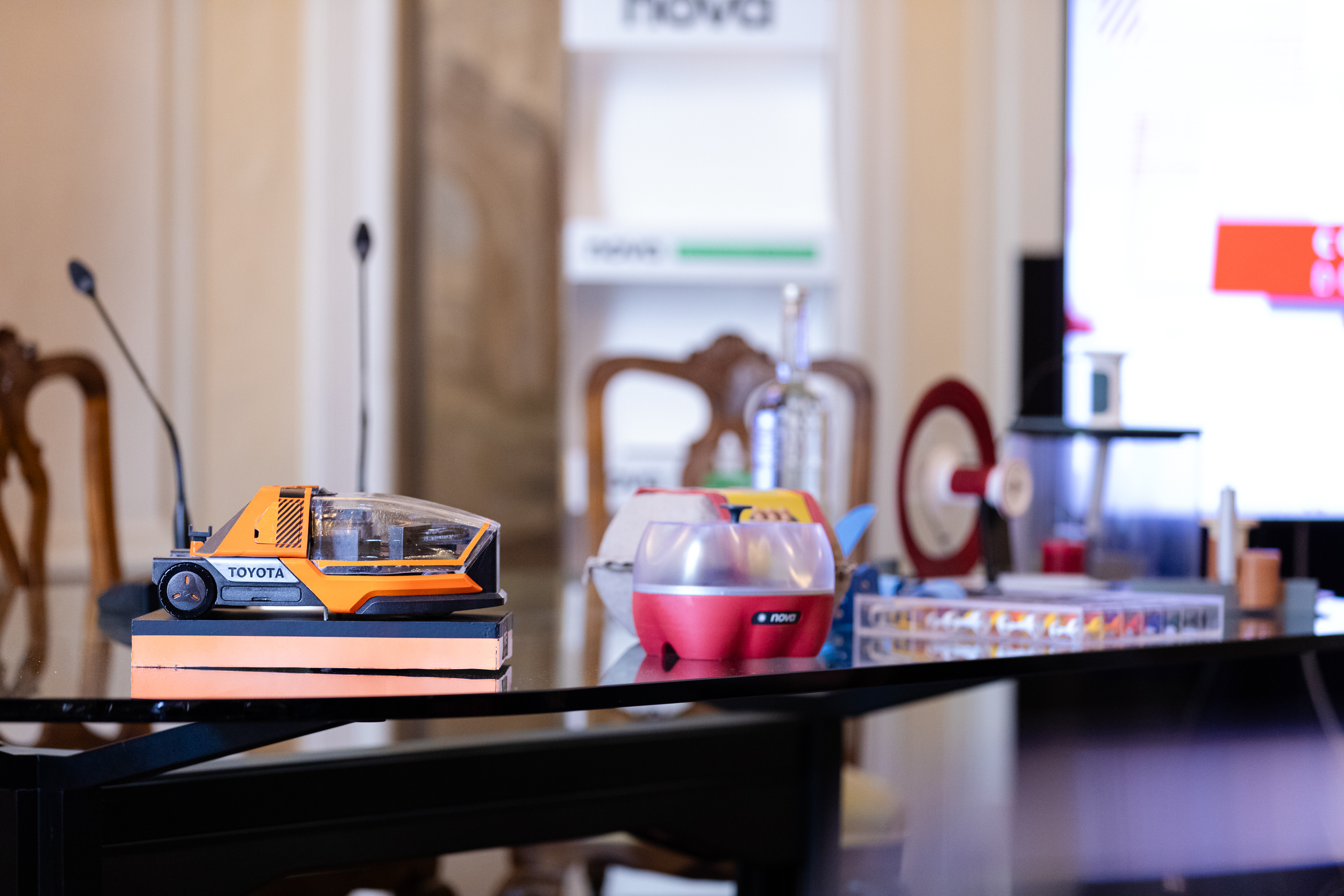 Modellino di auto Toyota arancione su un tavolo con oggetti vari e microfoni in lontananza.